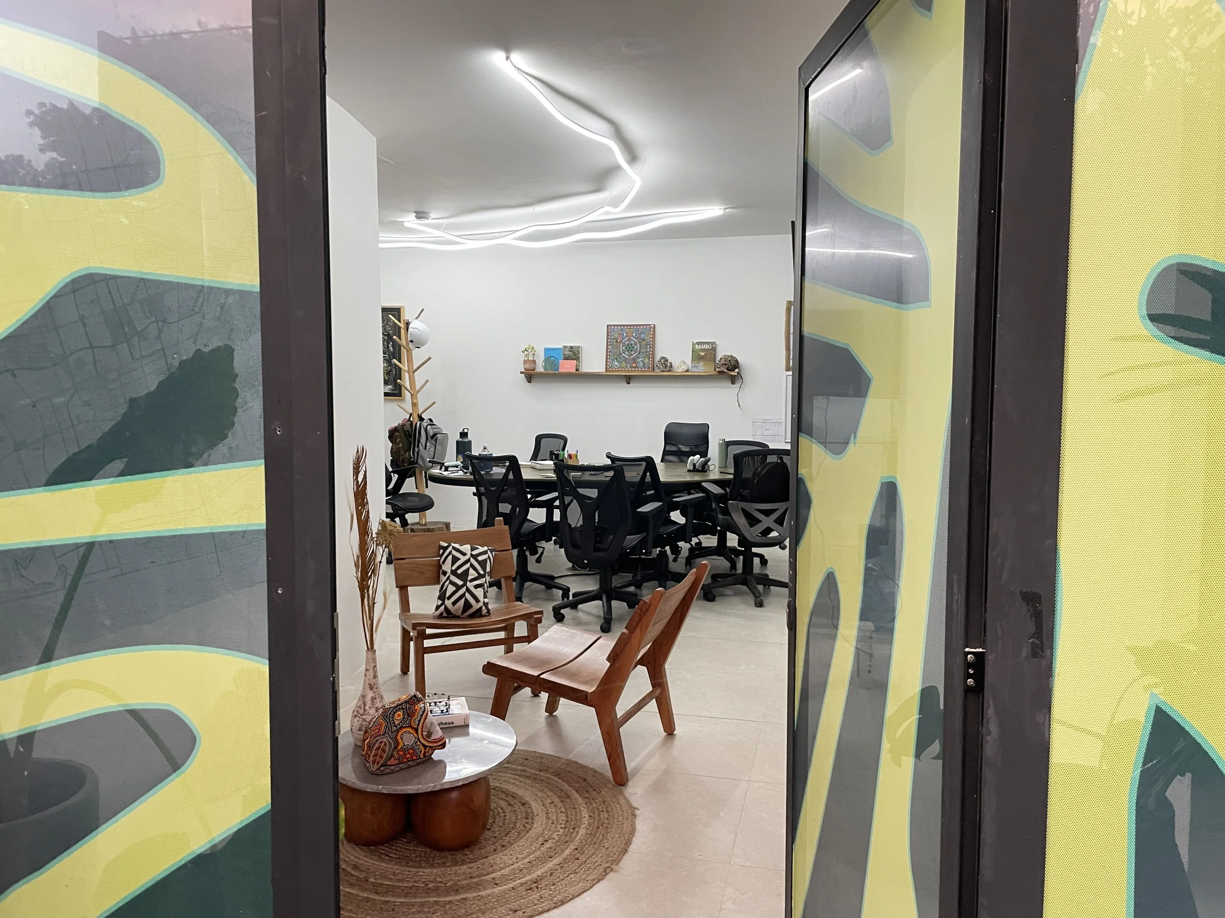 View of a modern office conference room with a circular table surrounded by black office chairs in the background. In the foreground, two wooden chairs with light-colored cushions are visible, along with a small round table holding a decorative bag a