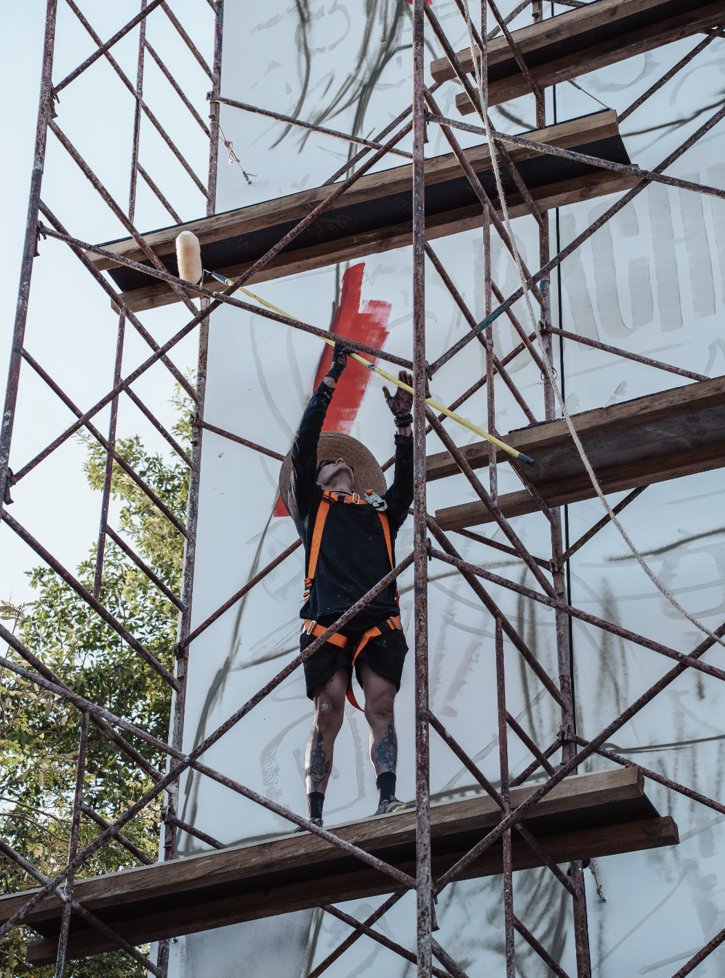A person painting a large white wall using a roller on an extension pole while standing on scaffolding.