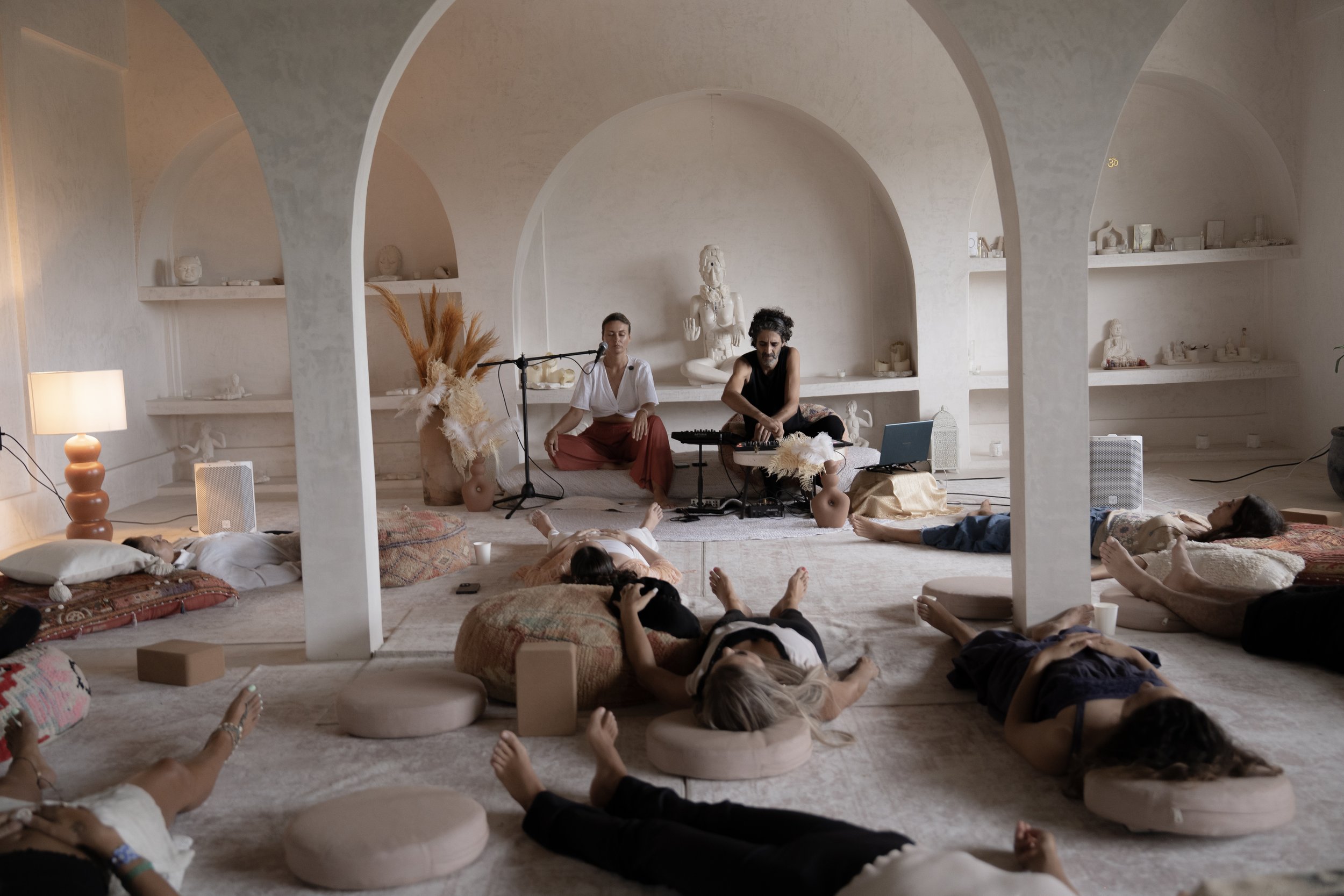 A group of people lying down on cushions and pillows on the floor of a cozy, arched, white-walled room with built-in shelves, some with small sculptures and decor items. In the background, two musicians are performing, one with a microphone and the o