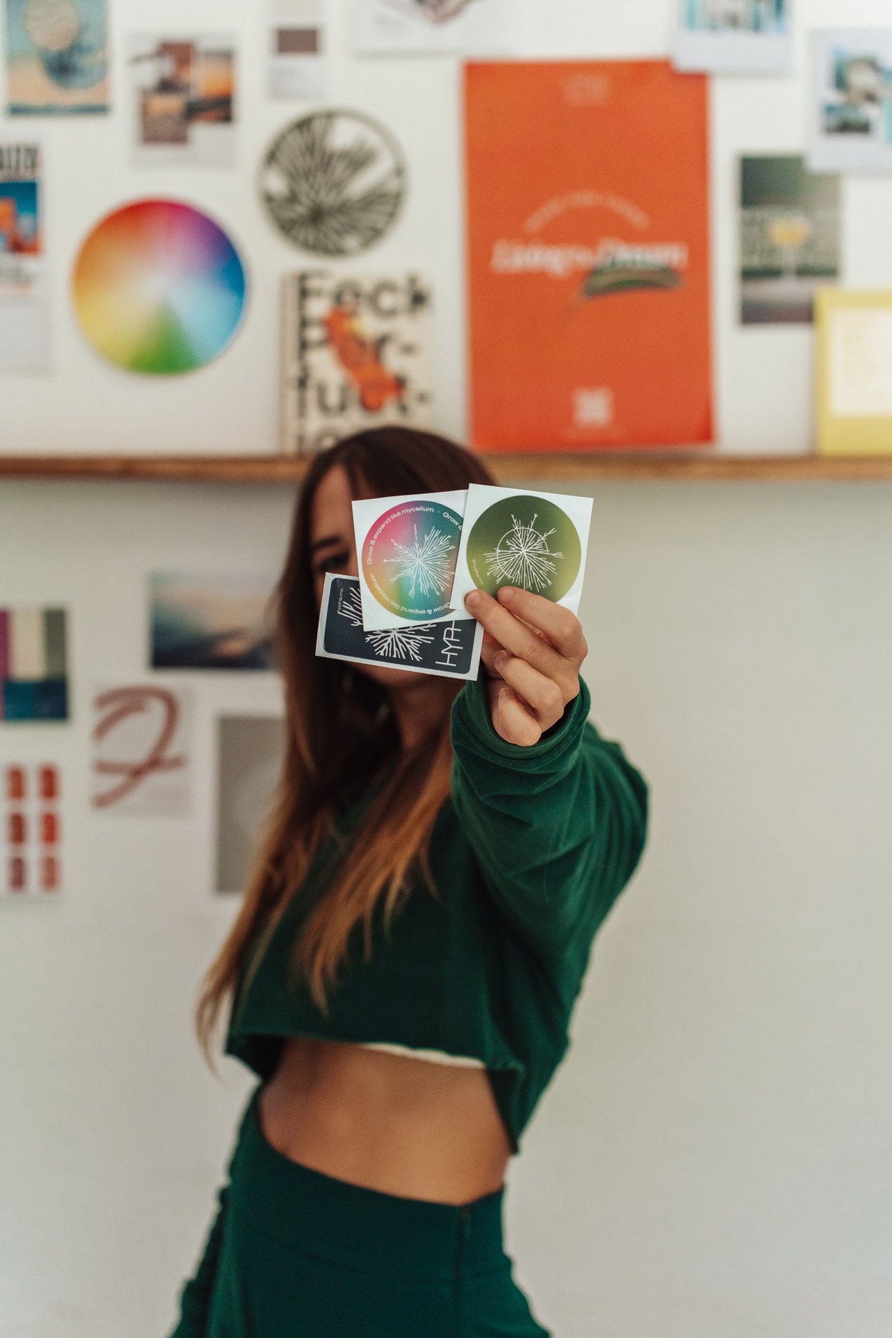 APOD Hypha with long hair holding several colorful stickers with a dandelion design in front of her in a creative workspace with posters and artwork on the wall.