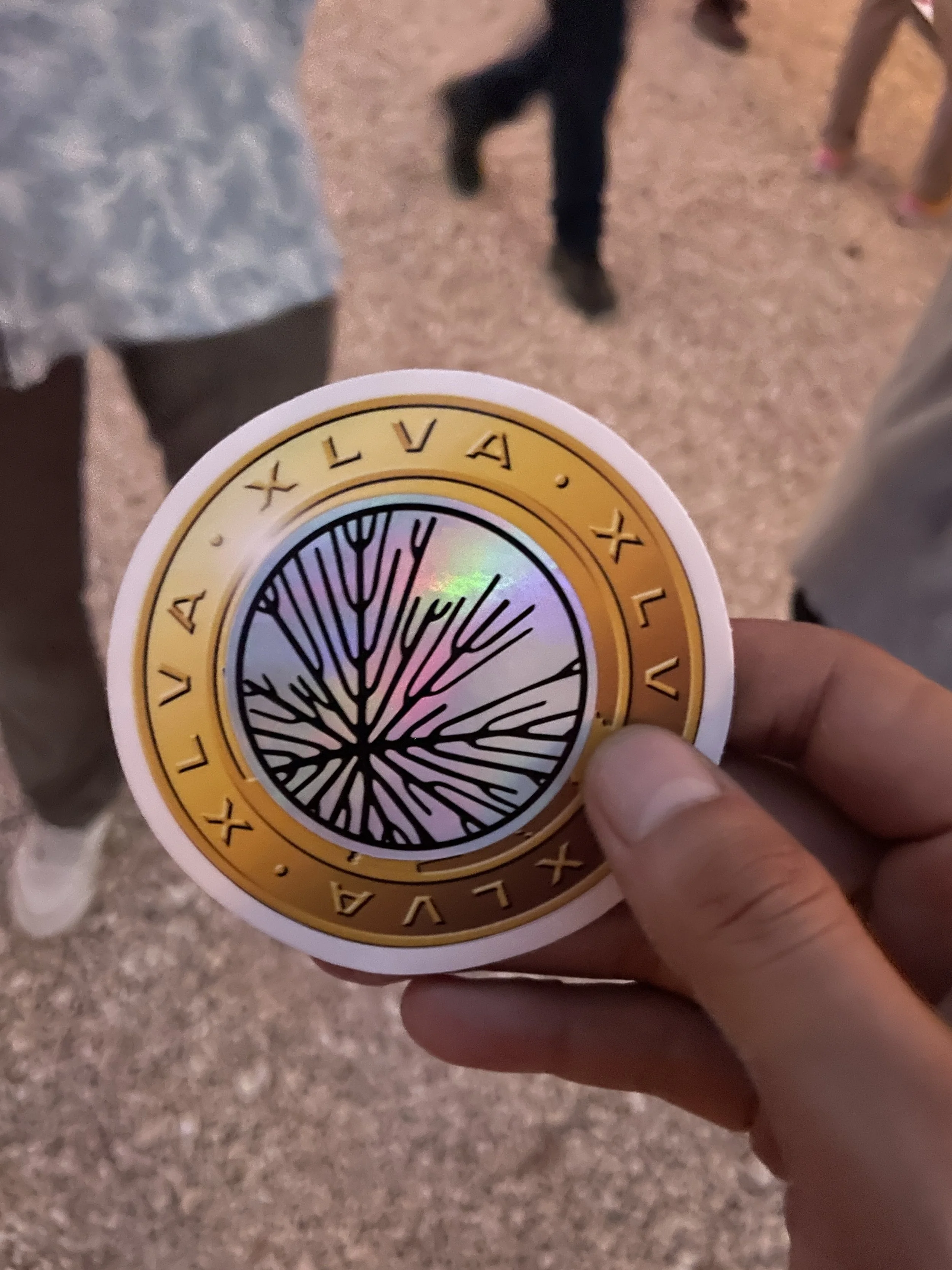 A hand holding a Hypha Inc. Xelva token with a tree design and a gold circular border. The background shows a ground surface and part of people's legs. Hypha Inc Marketing Agency and Creative Studio Lab, Tulum.