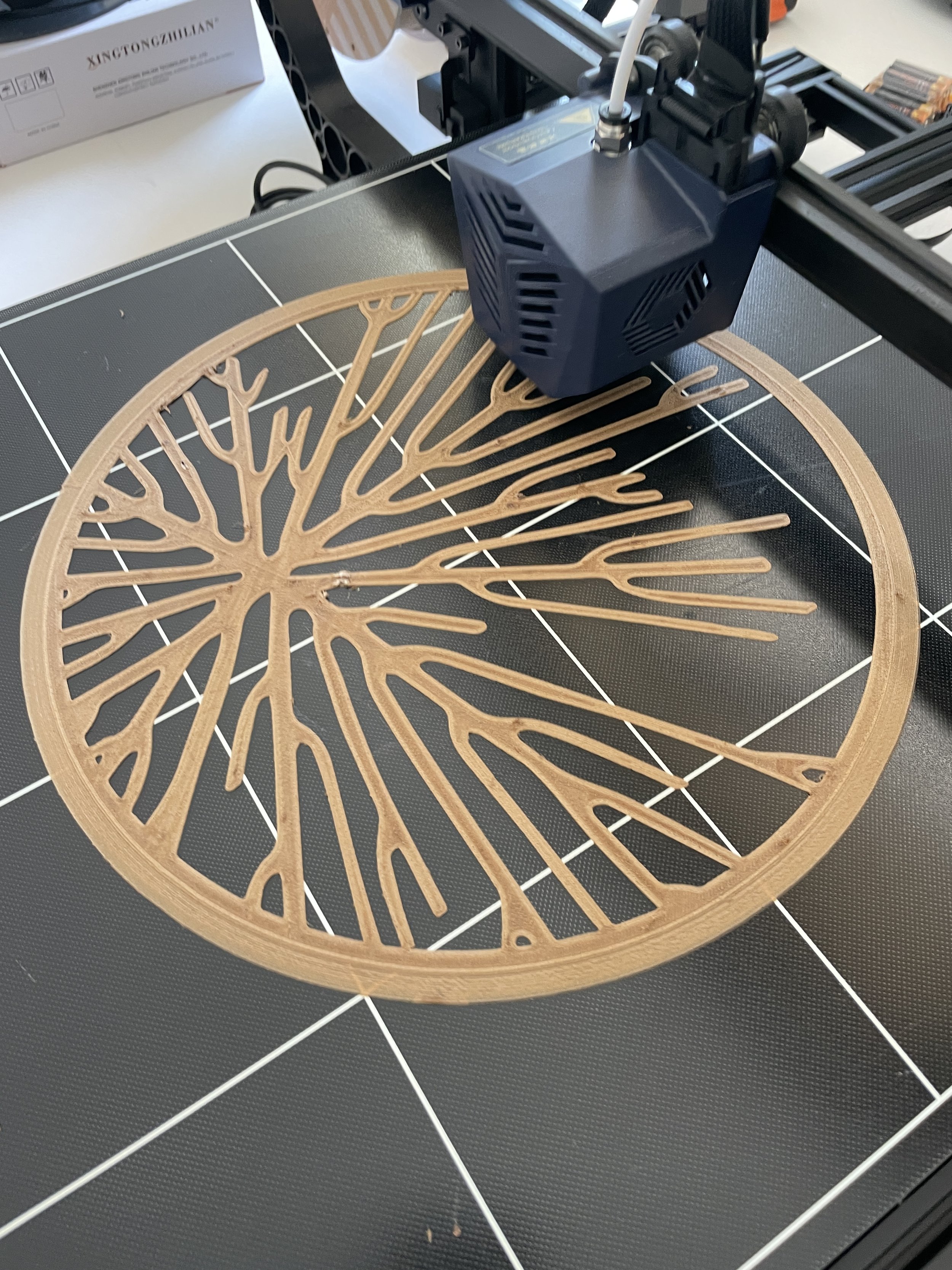 3D printer creating a circular, intricate tree-like design in beige filament on a black print bed. Hypha Inc. Logo