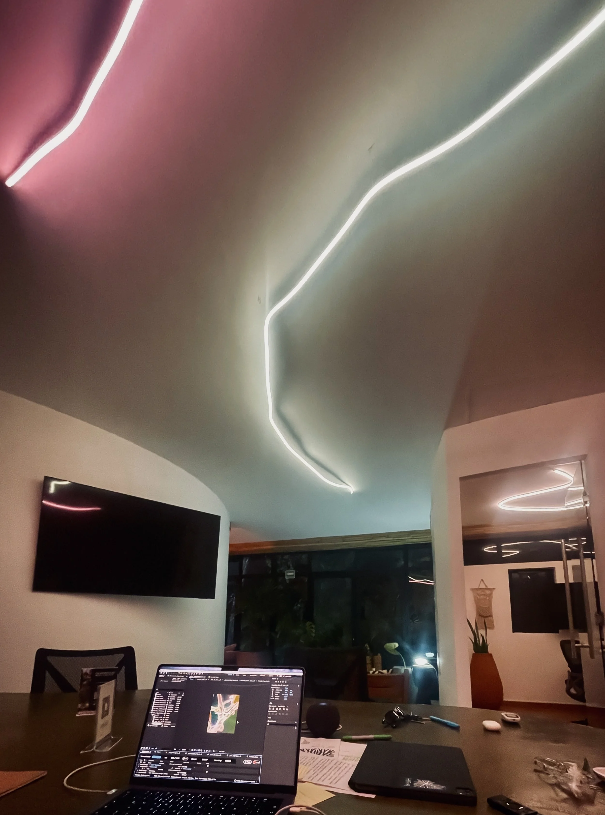Interior of a room with a modern ceiling featuring white LED strip lights, a wall-mounted flat-screen TV, a table with a laptop, papers, and various electronic devices, and large windows with outdoor greenery visible at night. Hypha Inc Marketing Age