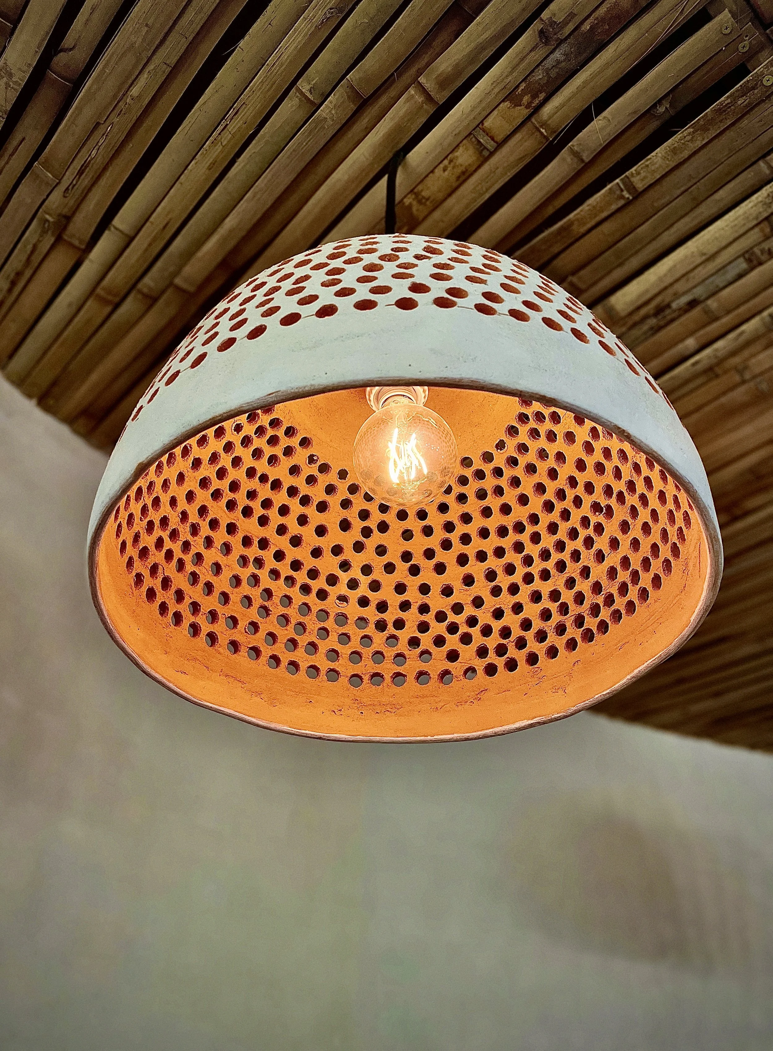Close-up of a modern ceiling light fixture with an orange perforated shade and incandescent bulb, mounted on a bamboo ceiling. Hypha Inc Marketing Agency and Creative Studio Lab, Tulum.