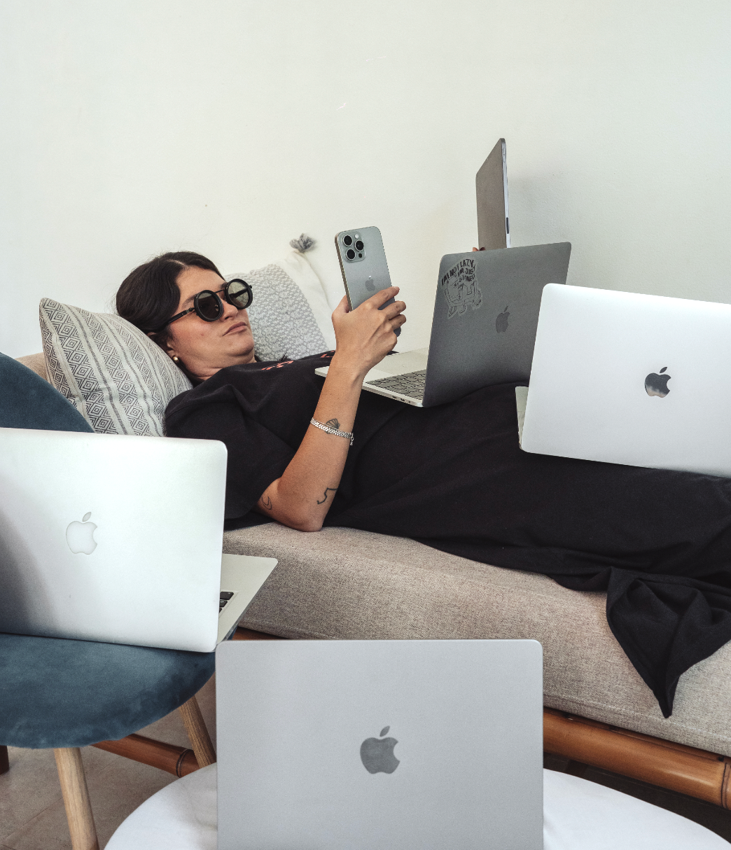 A woman with dark hair wearing sunglasses, lying on a sofa, surrounded by four Apple laptops. She is holding a smartphone in her right hand.