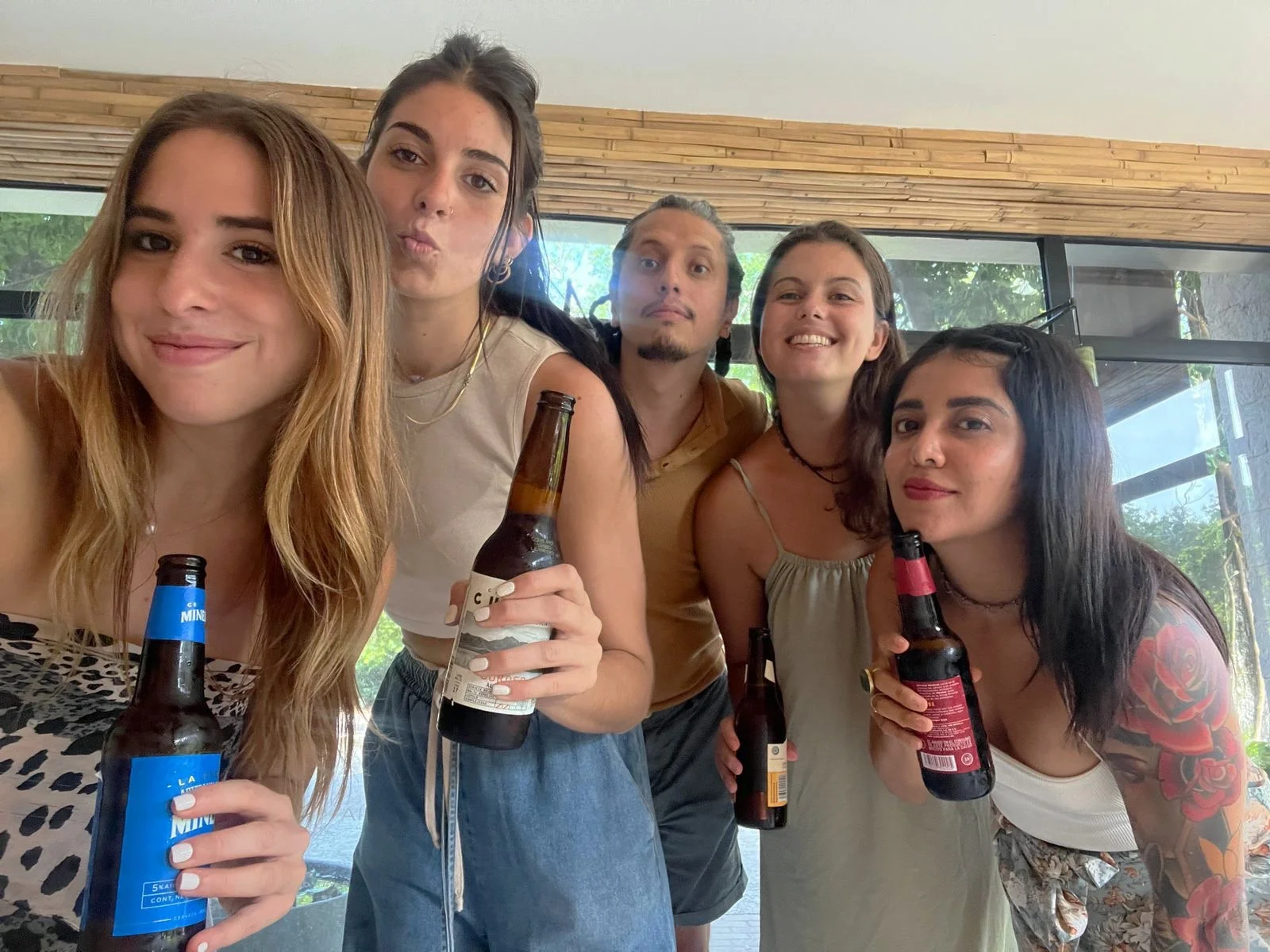 Group of five young adults smiling and holding beer bottles, taking a selfie indoors with large windows and greenery outside. Hypha Inc. Team Marketing Agency Tulum
