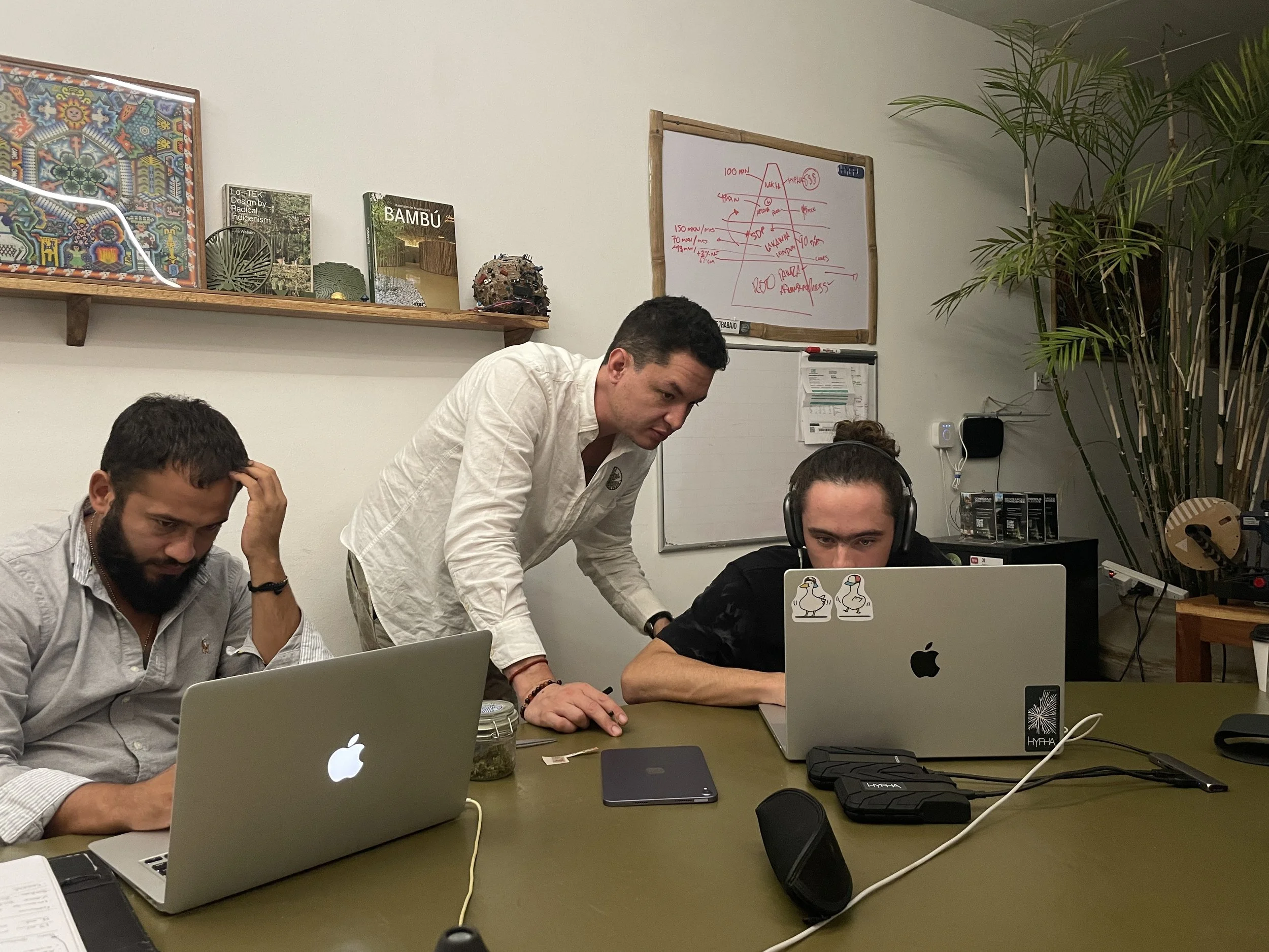 Three men working at a conference table with laptops; one man is standing and leaning over to look at a laptop of a seated man with headphones, while another man is seated with a laptop and appears to be thinking.