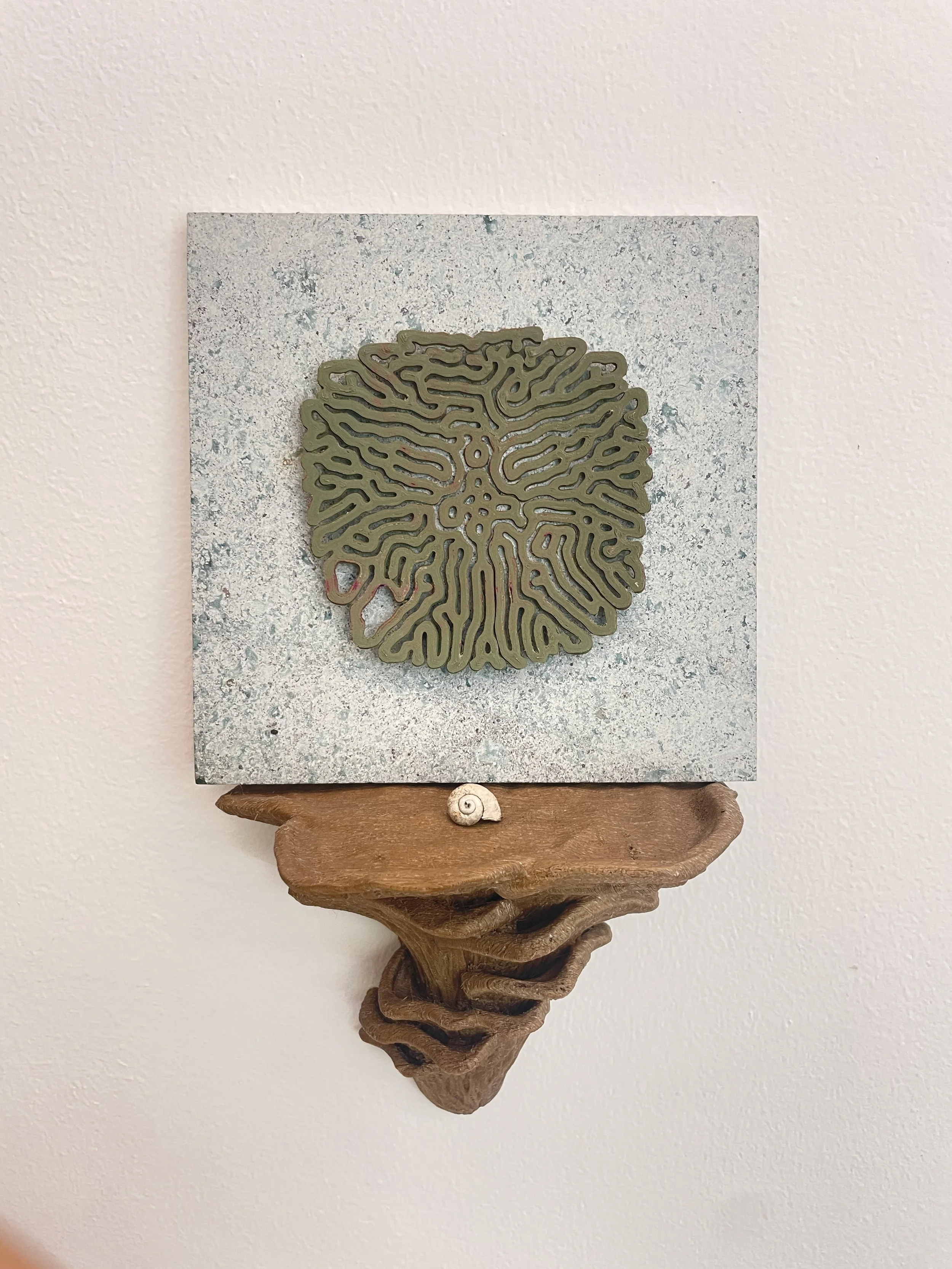 A piece of wall art featuring a matrix-like maze design on a square canvas, mounted on a wooden shelf with a small seashell decoration beneath. Hypha Inc Marketing Agency and Creative Studio Lab, Tulum.