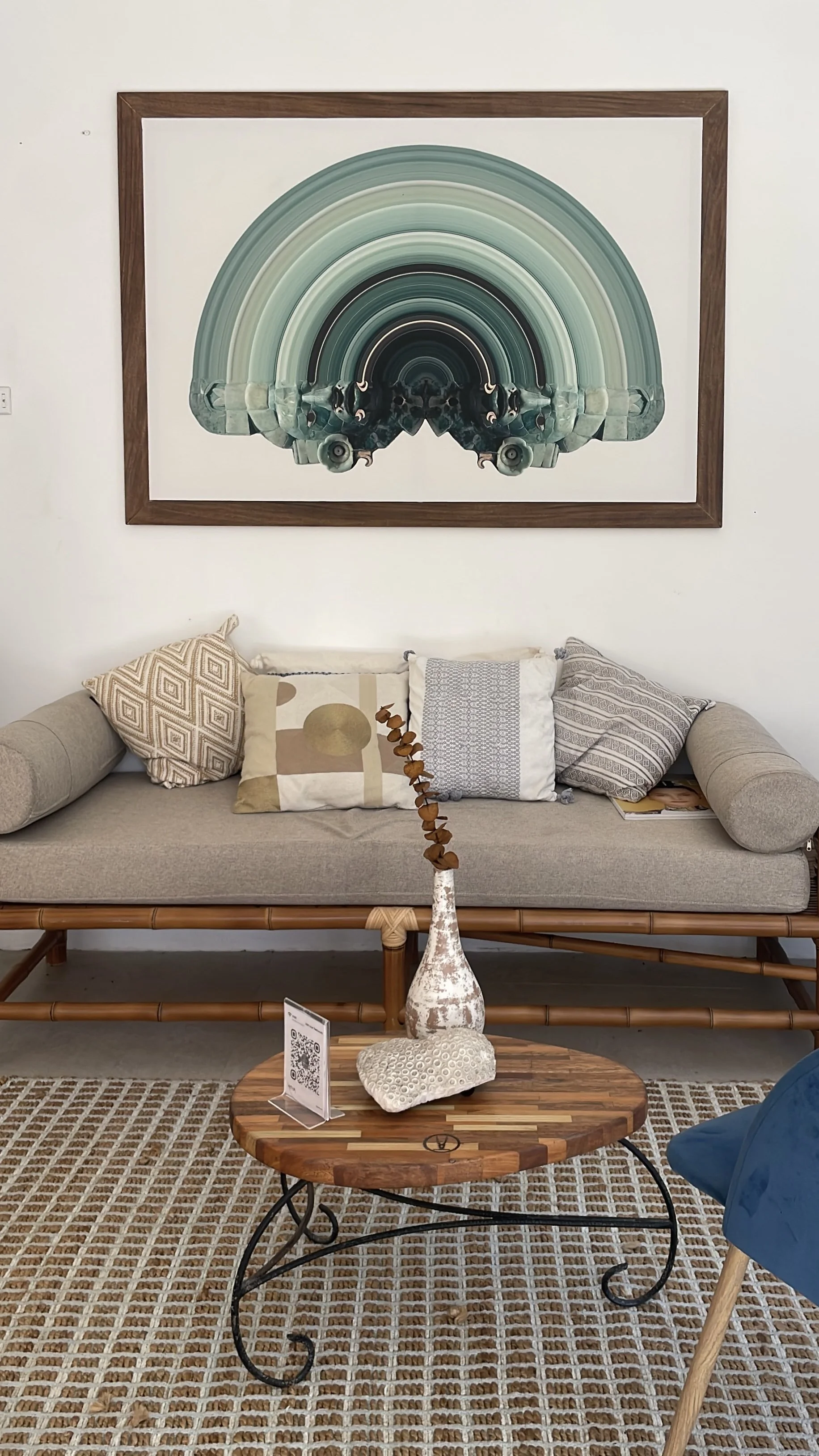 A living room with a beige sofa and throw pillows, a wooden coffee table with a decorative vase and a textured stone, a patterned rug, and a large framed abstract art piece with concentric arches in shades of blue and green on the wall. Hypha Inc Ads