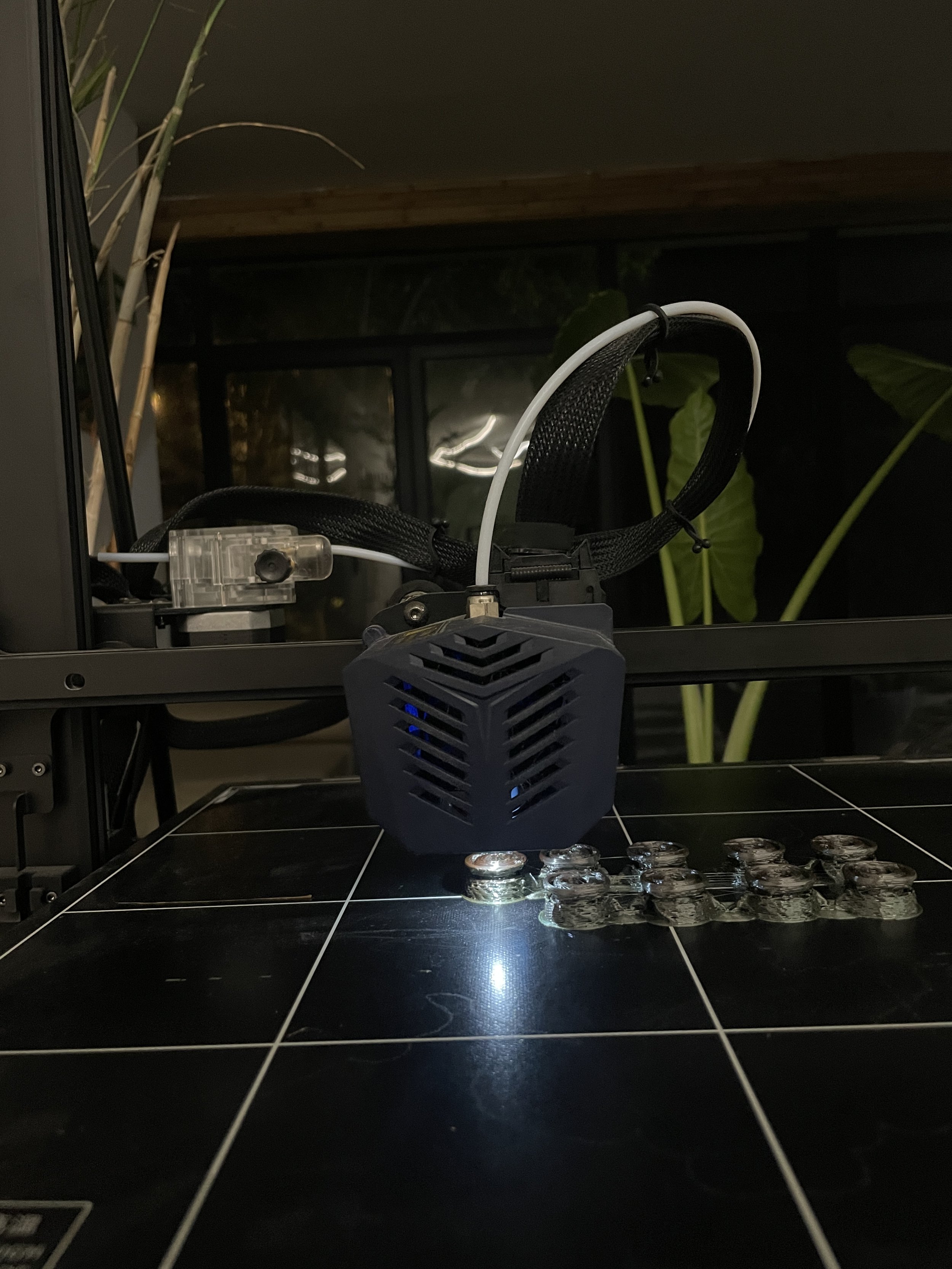 A 3D printer extruding filament onto a printed object on a black tiled surface in a dimly lit room with plants and a glass window in the background. Hypha Inc Marketing Agency and Creative Studio Lab, Tulum.