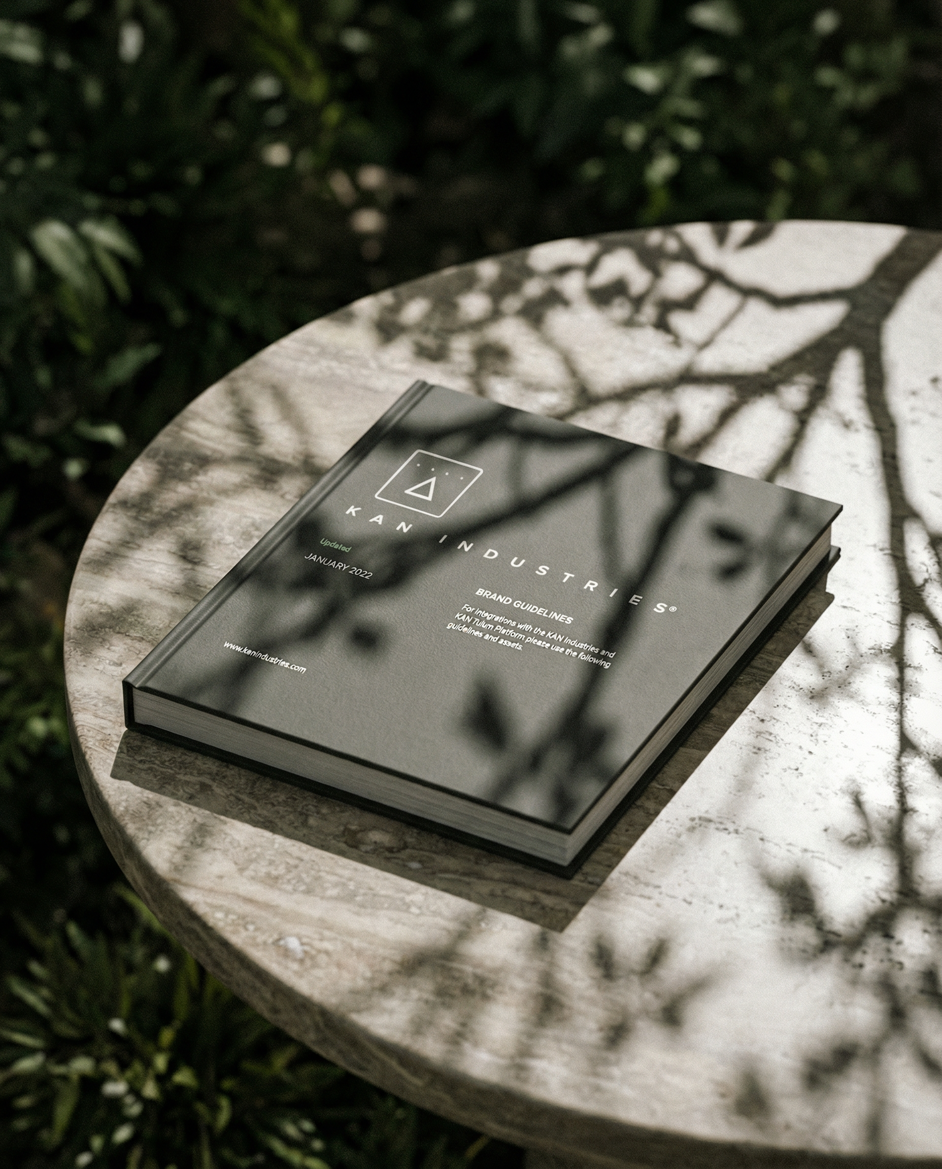 A black booklet titled 'KAN INDUSTRIES' resting on a round, light-colored stone table outdoors, with shadows of tree branches cast across the table and booklet. Hypha Inc. Branding and Design Agency Tulum