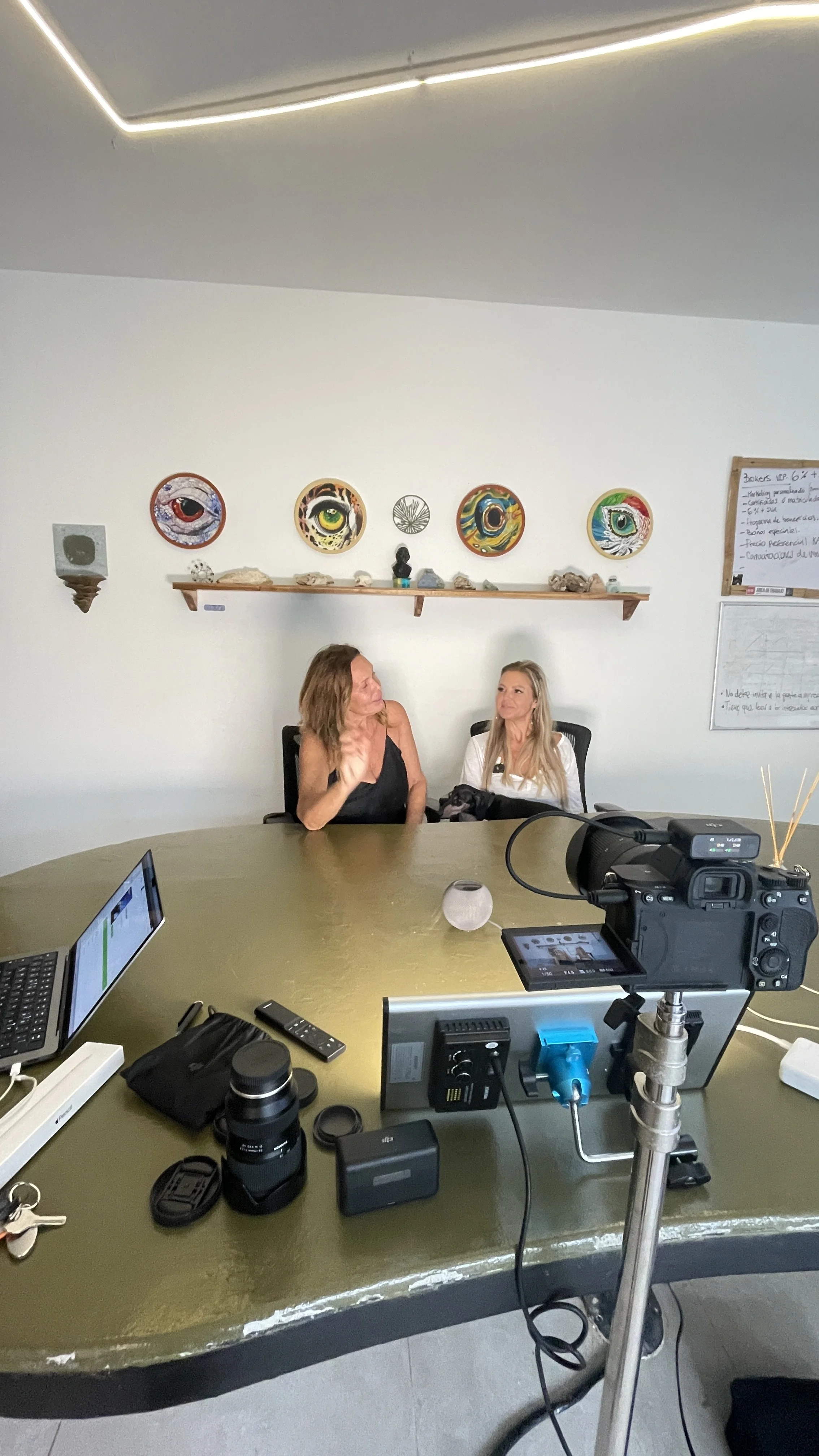 Two women are seated at a conference table in a room with white walls. One woman is speaking, and the other is listening. There is a camera on a tripod positioned to record the interview, along with laptops and other electronic devices on the table. 
