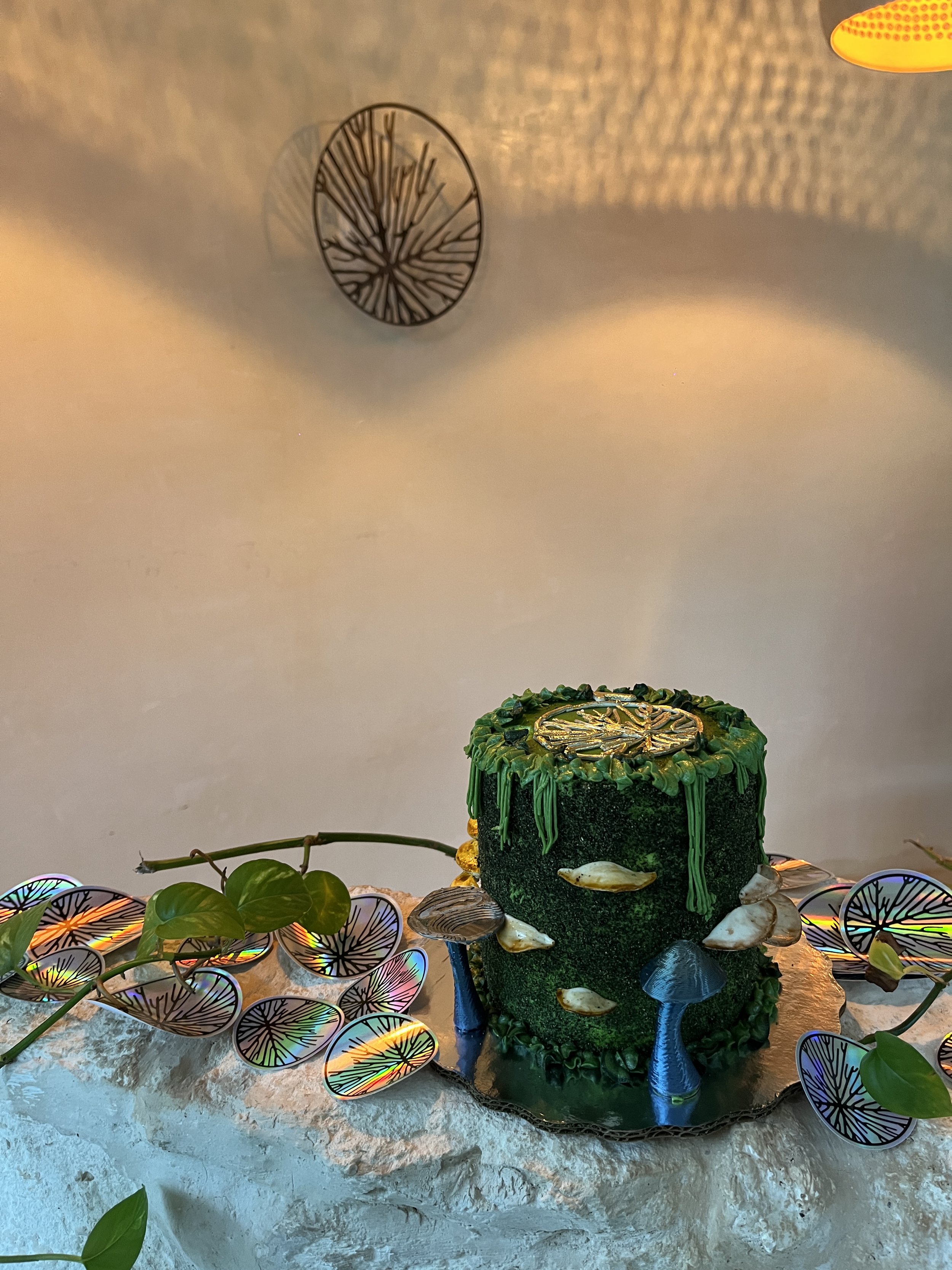 A cake with green and black colors decorated with mushrooms and a tree branch surrounded by holographic leaf decorations. The background has a wall with a shadow pattern and a circular wall decor. Hypha Inc Marketing Agency and Creative Studio Lab
