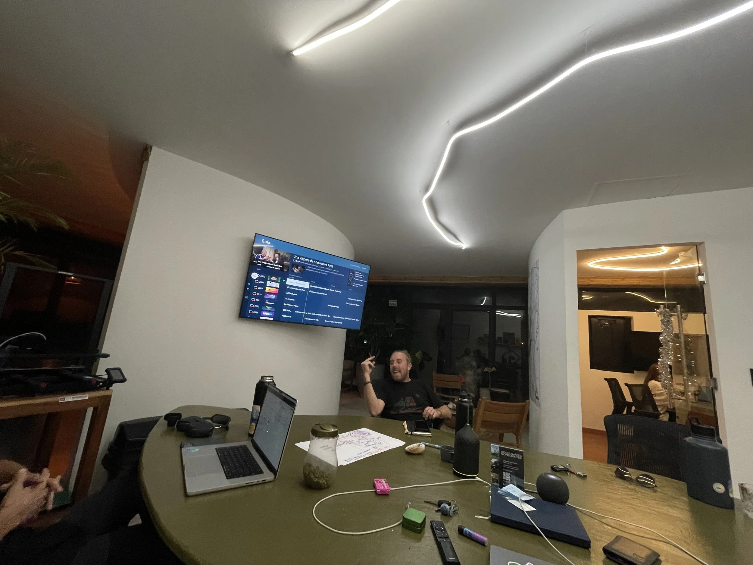 A person sitting at a table in a modern room with a curved ceiling and LED lighting, watching a television mounted on a white wall.