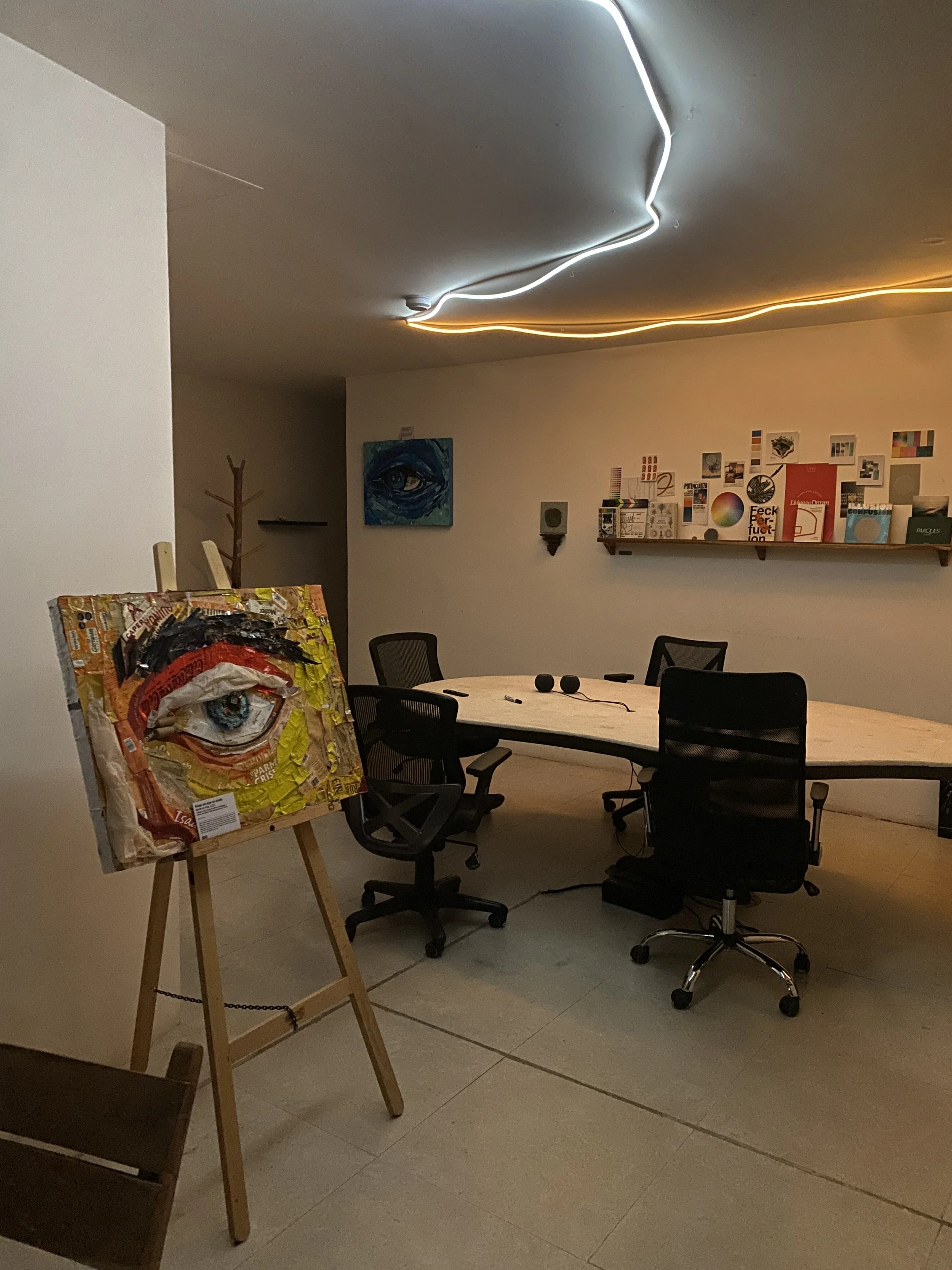 An art gallery or creative workspace with a large oval table surrounded by office chairs, a colorful collage painting on an easel featuring an eye, wall-mounted artwork, and illuminated, wavy ceiling lights. Hypha Inc.