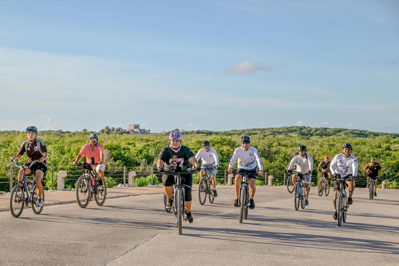 Group of cyclists riding bicycles outdoors on a clear day with green foliage and blue sky in the background in Parque del Jaguar Tulum
