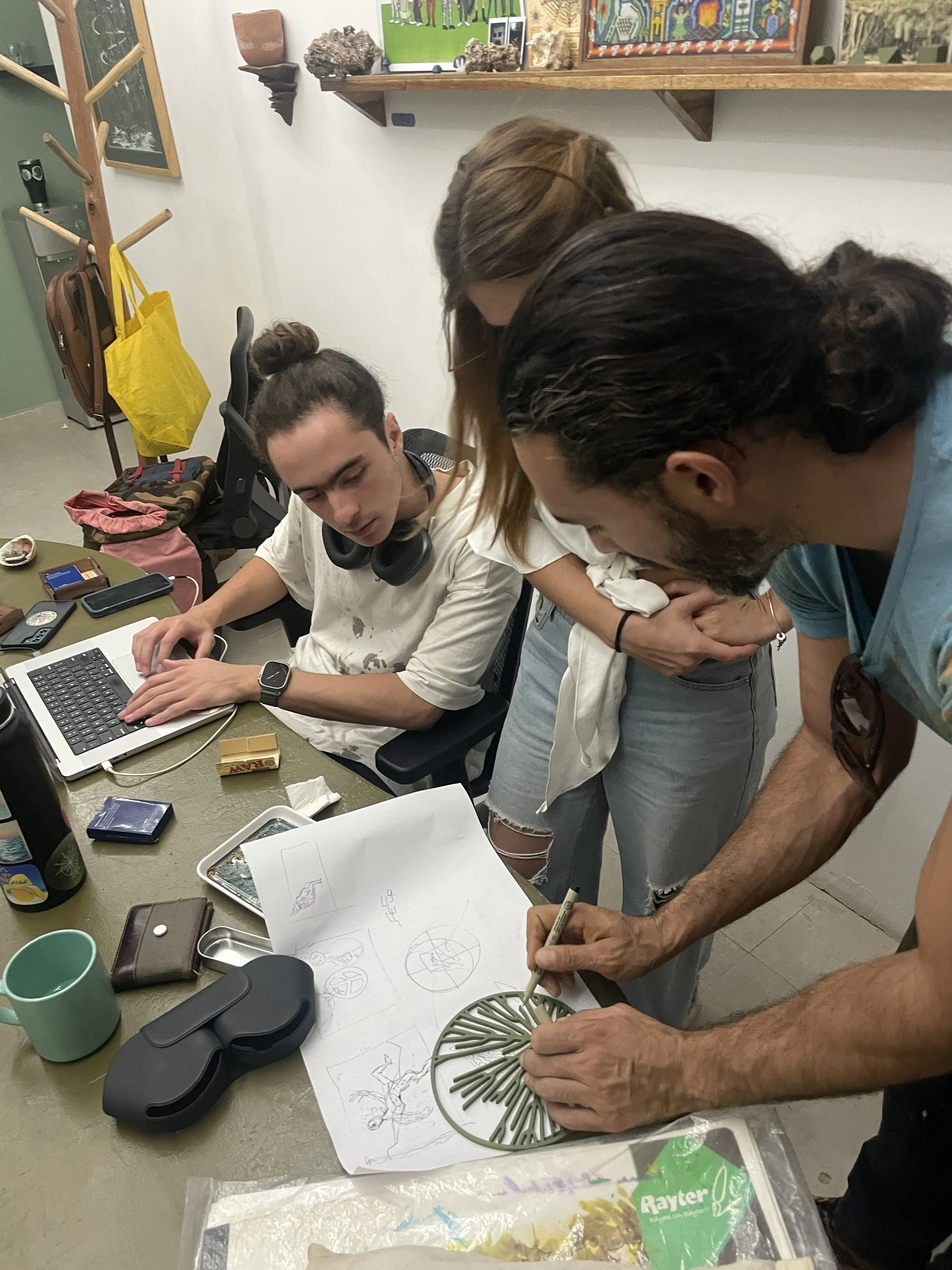 Group of three people gathered around a table with various items, with one person drawing on a circular object with a marker, while the others observe. The table has a laptop, notebooks, a mask, and a mug.