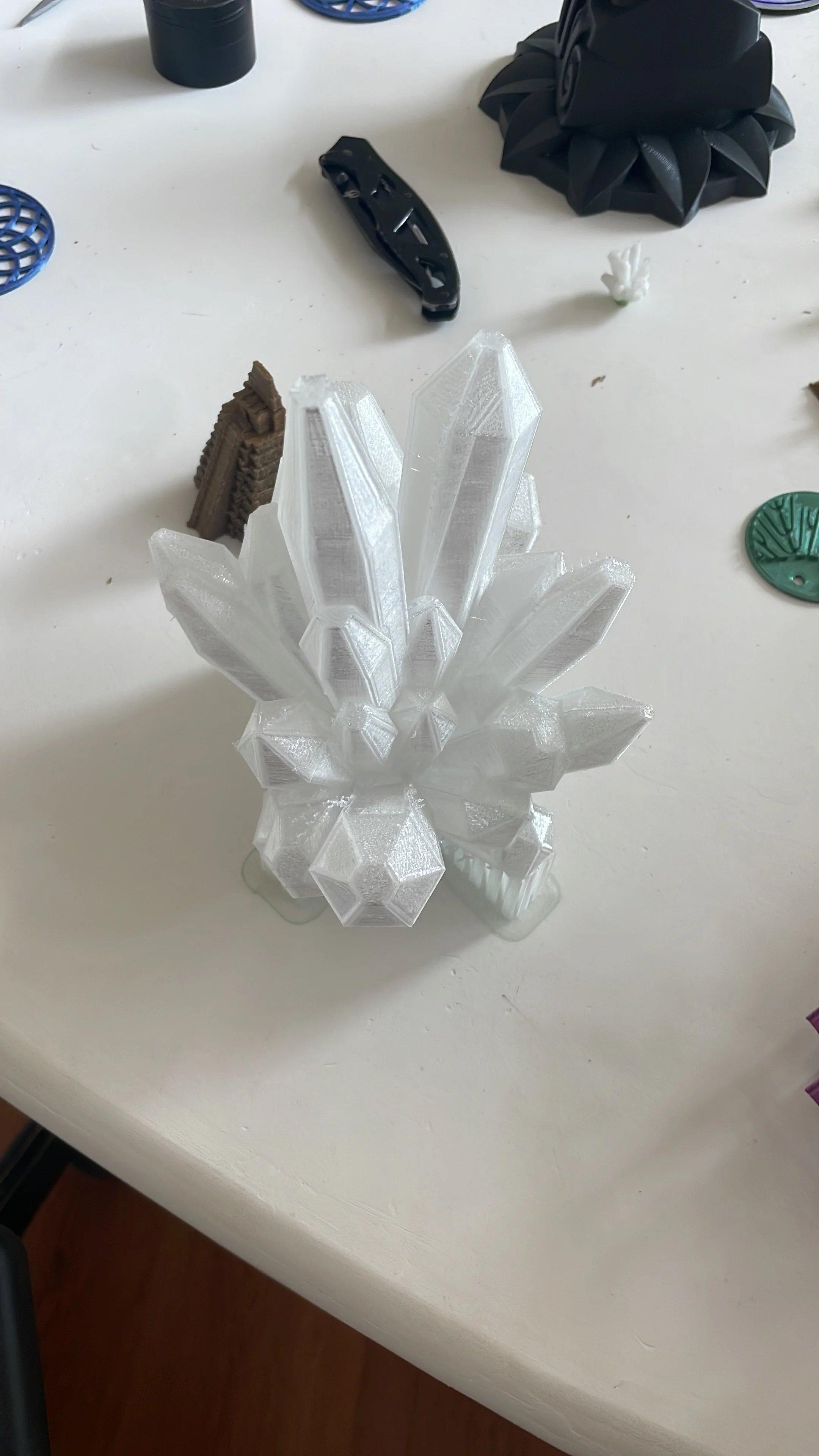 3D printed crystal cluster model on a white surface with miscellaneous objects around, including a black geometric object, a small white sculptural piece, a green token, and a black clip. Hypha Inc Marketing Agency and Creative Studio Lab, Tulum.