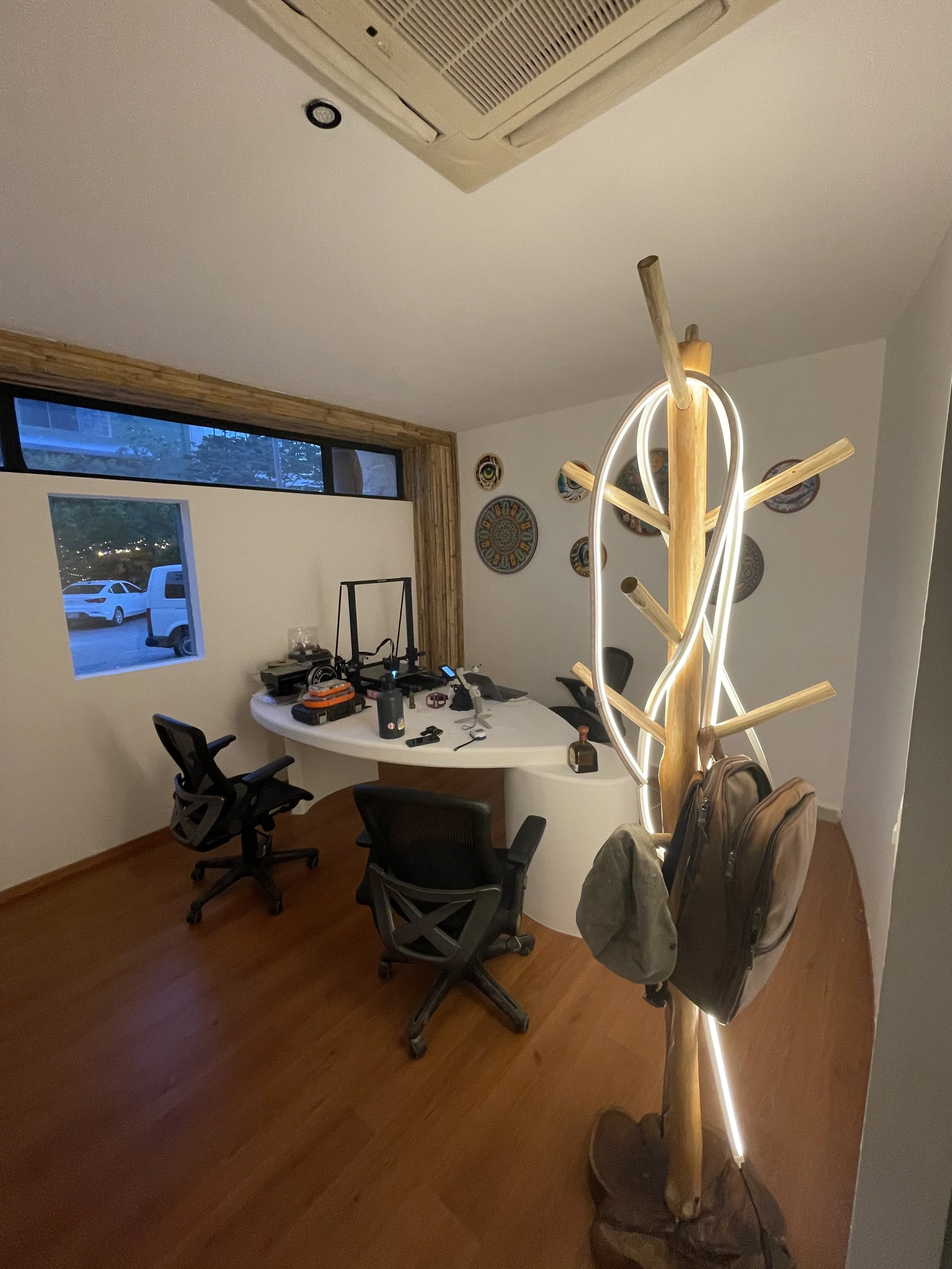 An office with a white desk, three black office chairs, decorative plates on the wall, a large window, and a wooden tree-shaped coat rack with LED lighting, backpacks, and hats hanging on it.