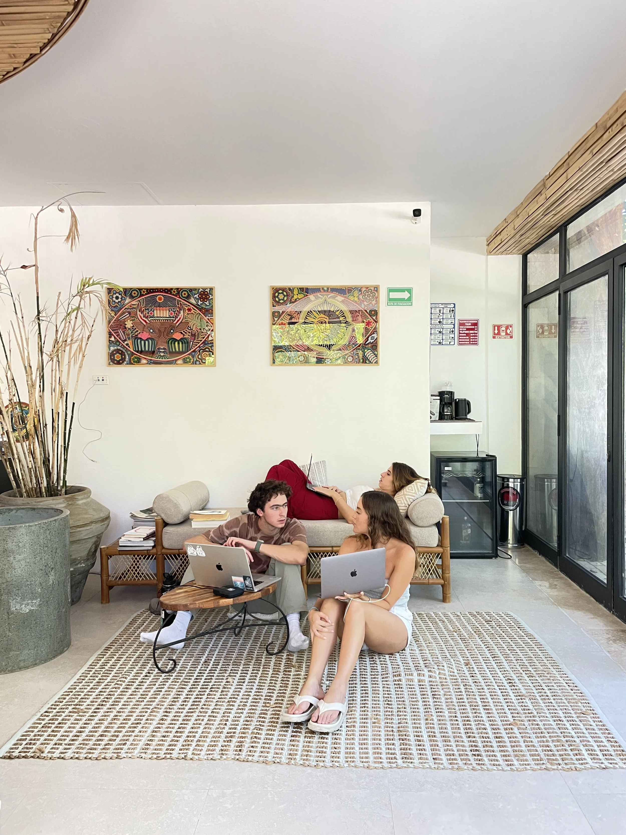 Three people in a modern, bright lobby or waiting area with white walls and artwork. Two are sitting on the floor with laptops, and one person is lying on a sofa reading.