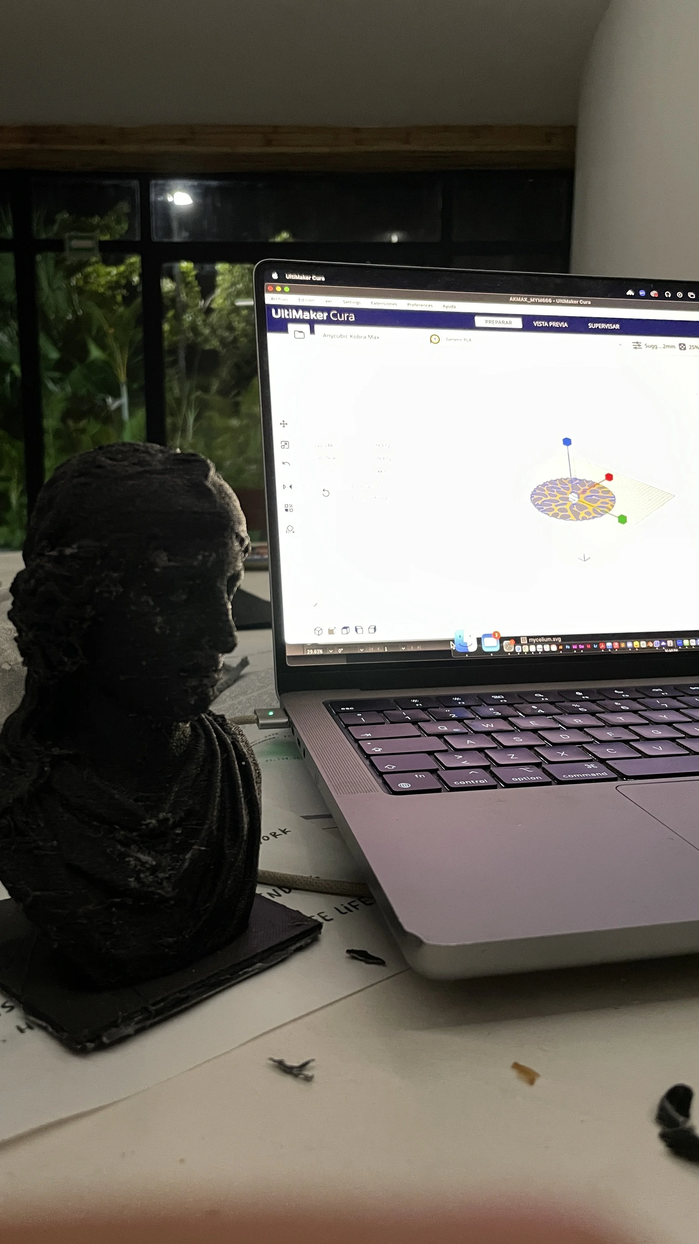 A laptop on a desk displaying 3D printing software, with a small bust sculpture next to it, and a window showing greenery outside in the background. Hypha Inc Marketing Agency and Creative Studio Lab, Tulum.