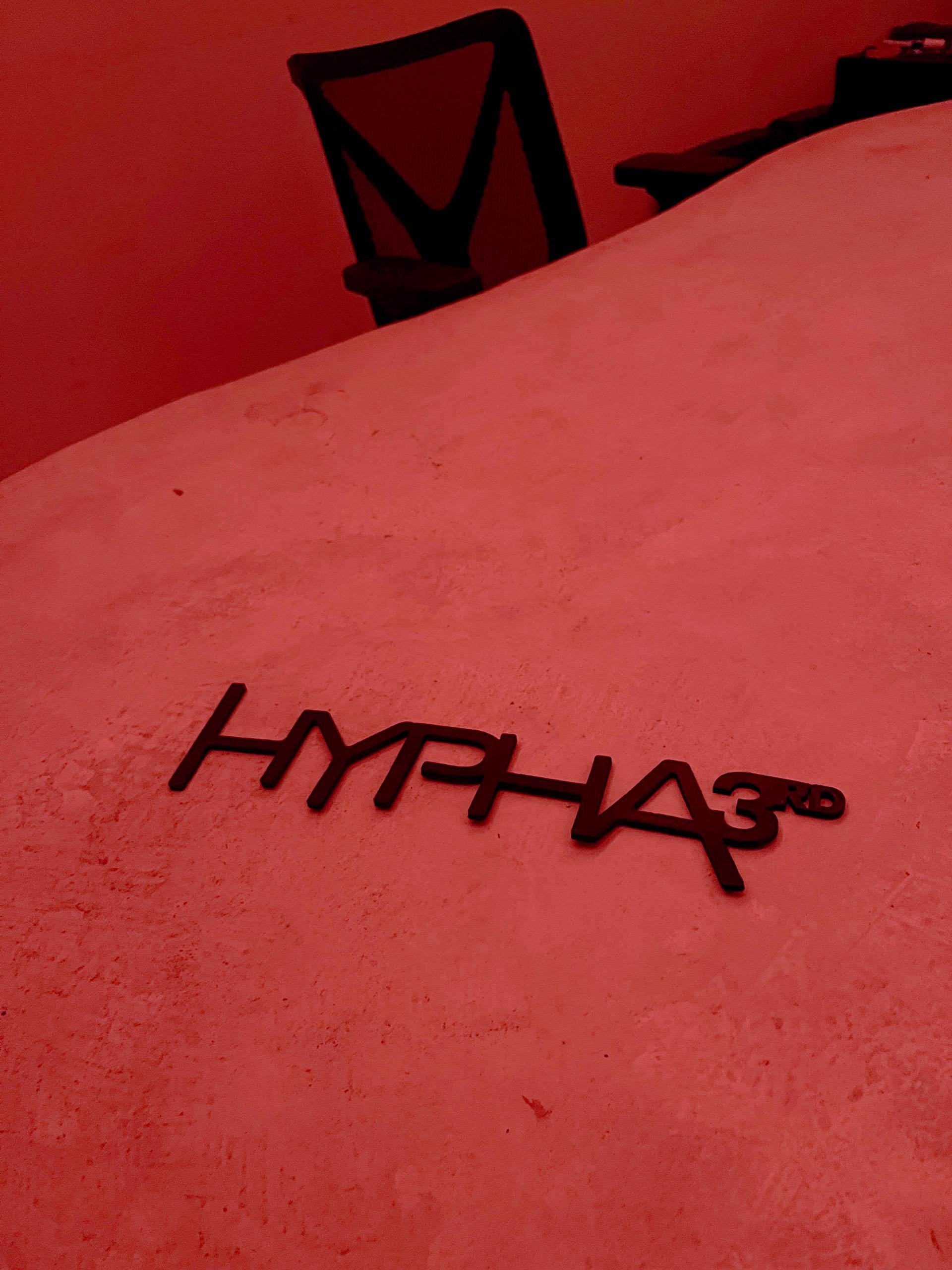 A black Hypha 3rd Anniversay logo on a textured pinkish surface with a chair in the background.