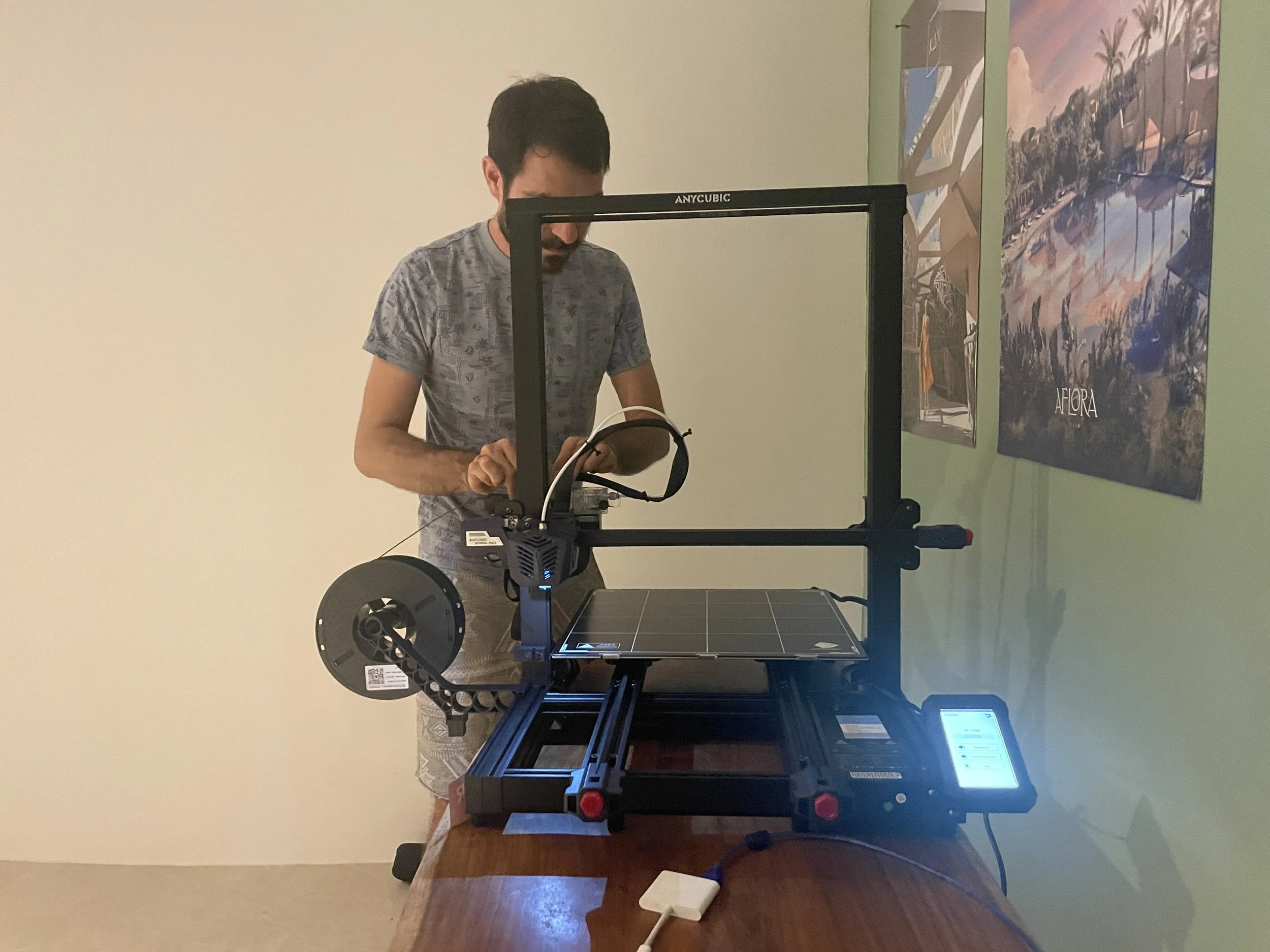 A man is standing at a wooden table using a 3D printer, which is in the process of printing an object. The printer is on, and the man is operating it while looking at a device in his hands. There are two colorful posters on the green wall to his righ