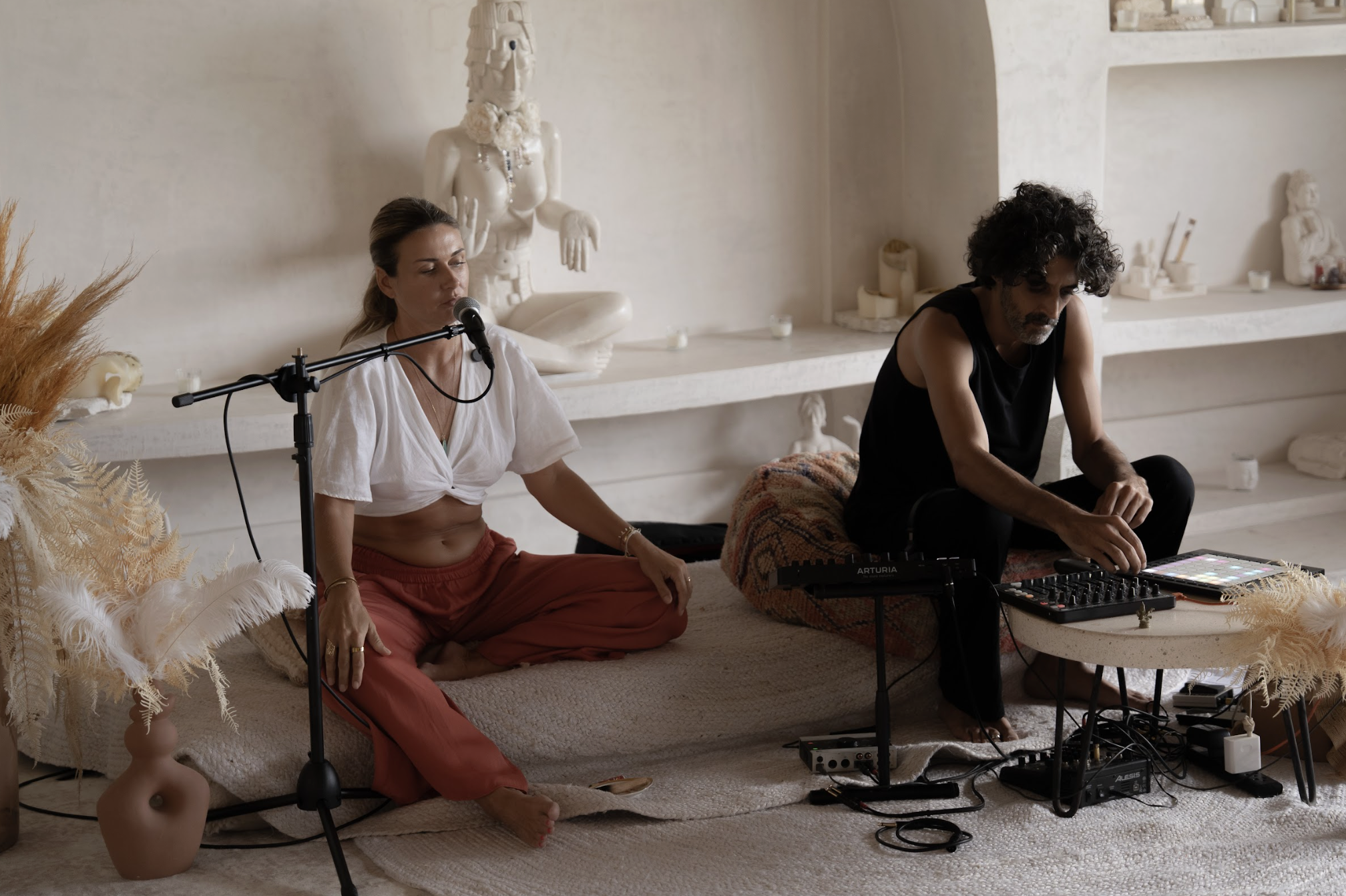A woman meditating with a microphone nearby and a man using electronic music equipment in a minimalist, bohemian style room decorated with white statues and dried plants.