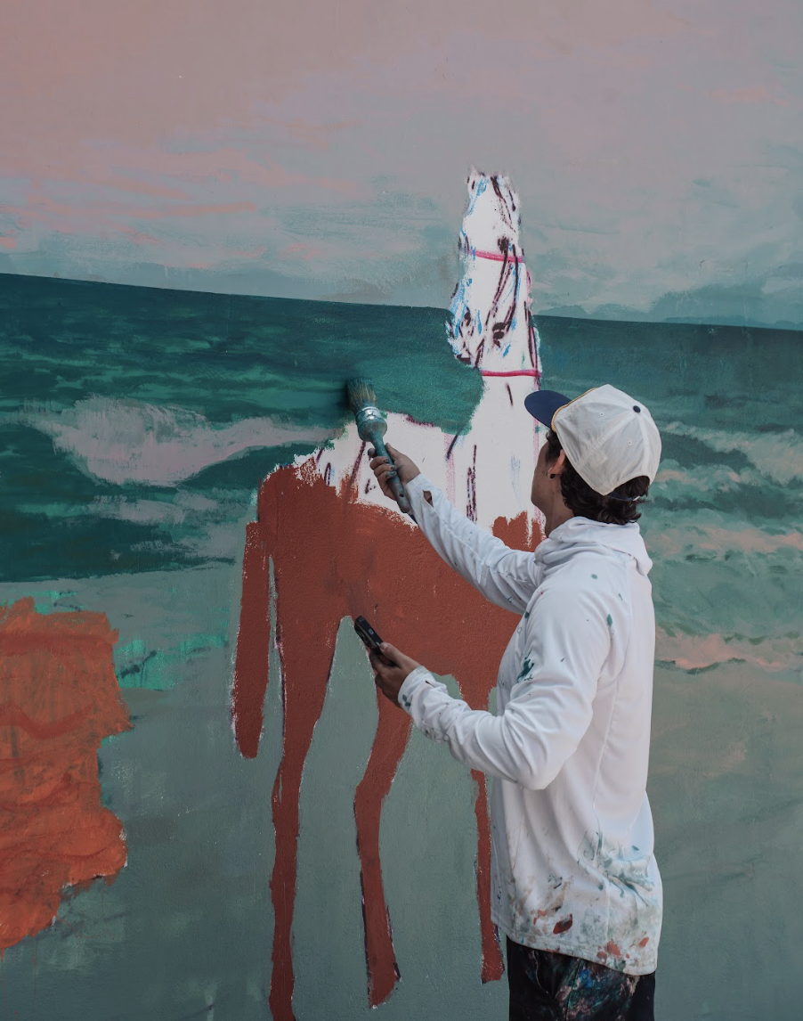 Person painting a large mural of a horse on a wall with ocean scenery in the background. The person is wearing a white jacket with paint stains, a white cap, and is holding a paintbrush and a phone.