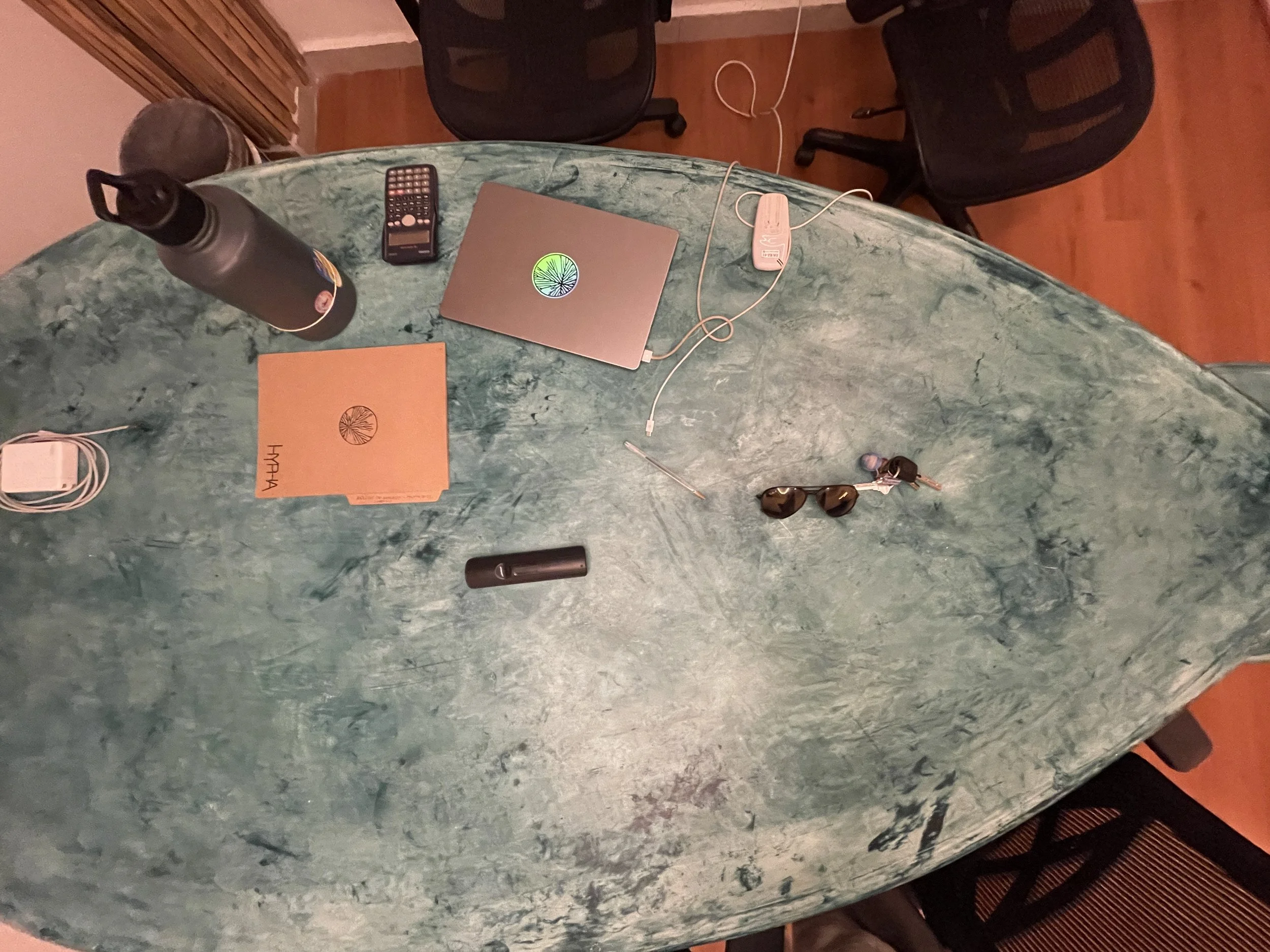 Overhead view of a green marble table with various items: a black water bottle, remote control, closed laptop with a sticker, open laptop with a sticker, white charging cable, black cylindrical object, sunglasses, and a set of keys with a keychain.