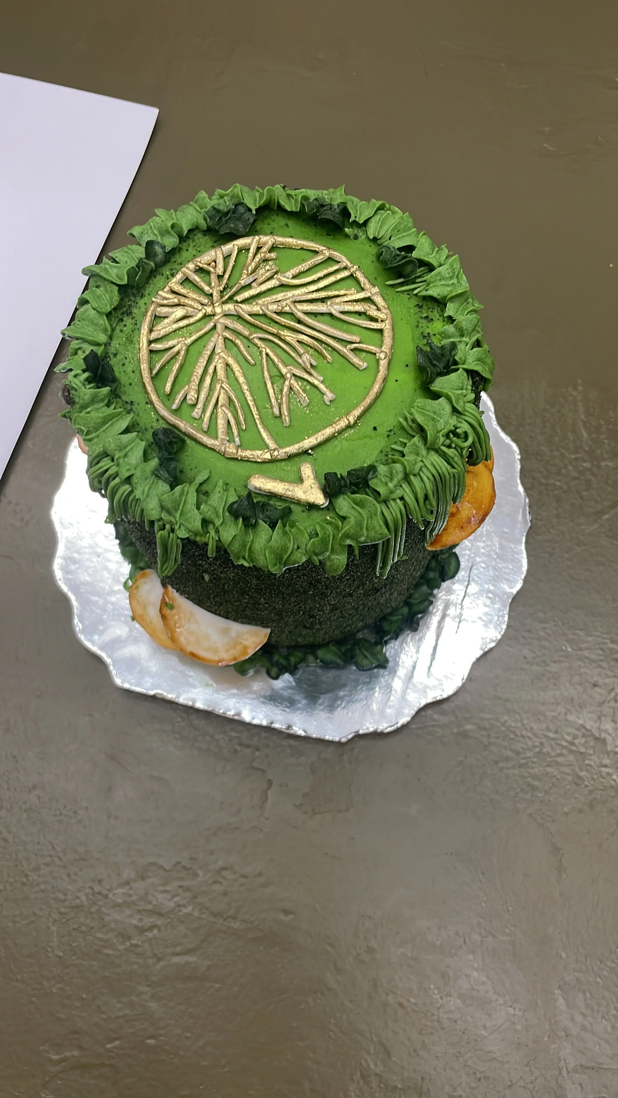A round cake with green frosting, decorated with black and green accents, featuring a hypha inc. logo and a gold arrow pointing downward on top. Hypha Inc Marketing Agency and Creative Studio Lab, Tulum.
