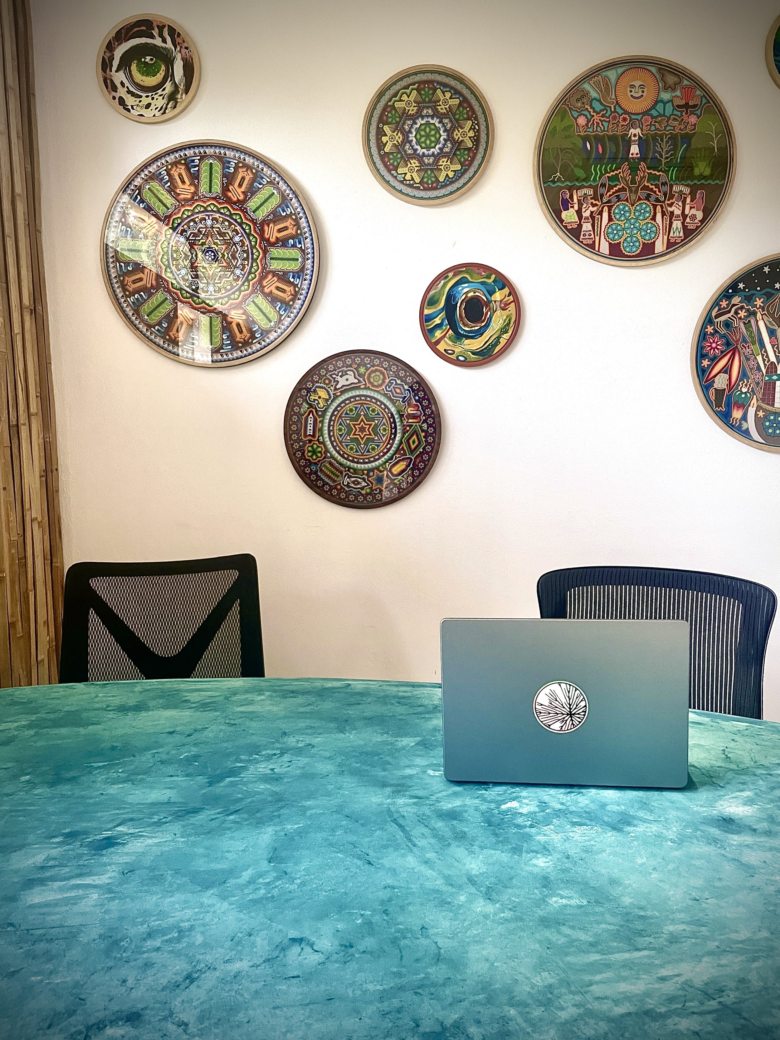 Office conference room with a turquoise table, two chairs, and a laptop with a tree logo, decorated with colorful circular art on the wall. Hypha Inc Marketing Agency and Creative Studio Lab, Tulum.