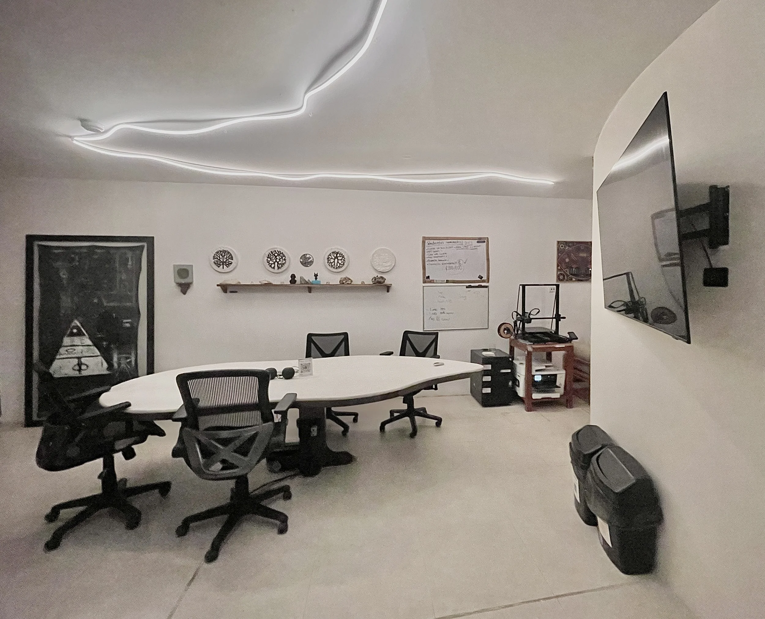 An office conference room with a white, irregularly shaped table surrounded by black mesh office chairs. The wall features a shelf with decorative plates and small statues, and whiteboards with notes. A large flat-screen TV is mounted on the right wa