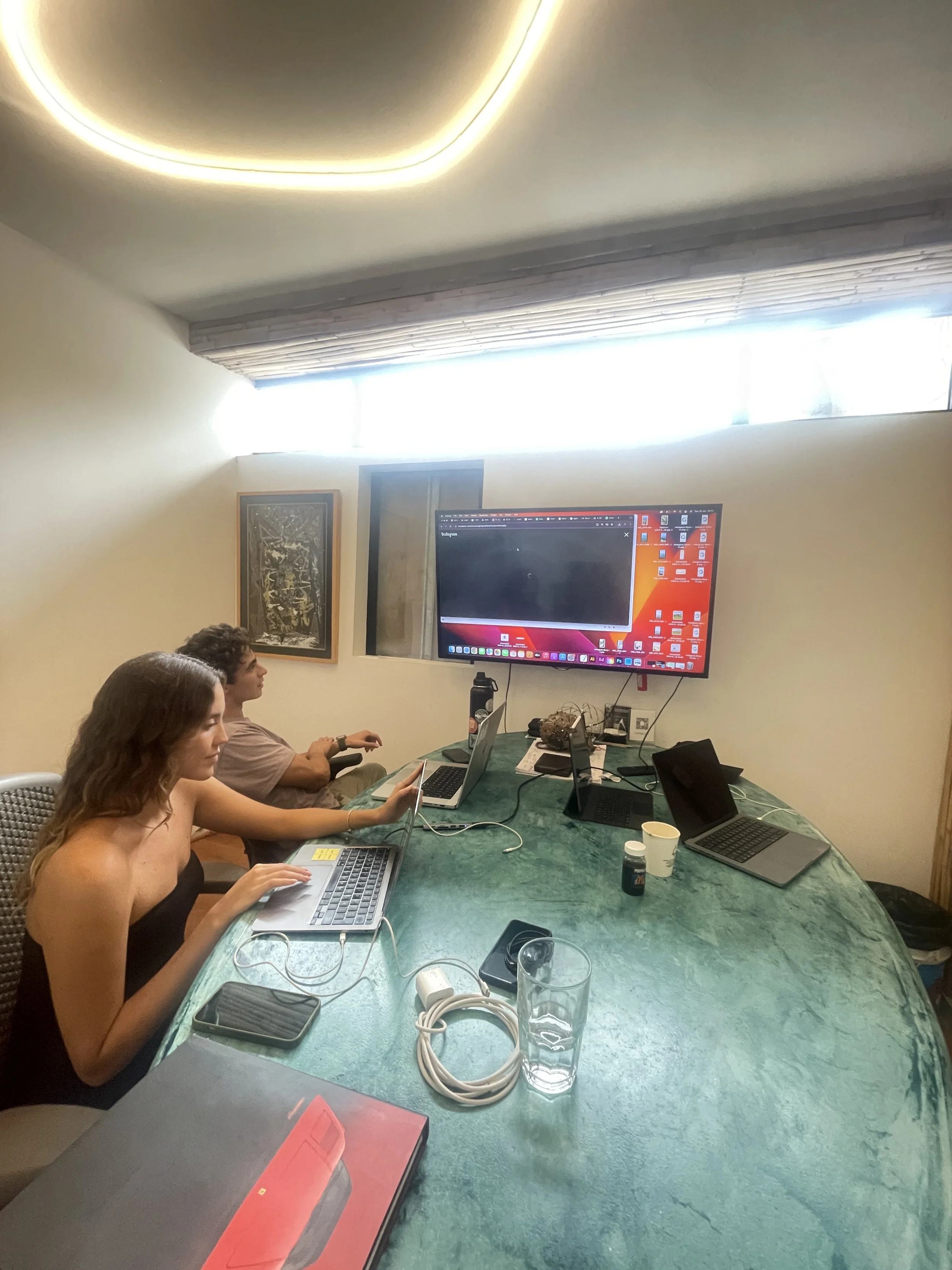 Two people sitting at a green marble conference table using laptops, in a room with a large monitor, a window with blinds, and various electronic devices and accessories on the table.