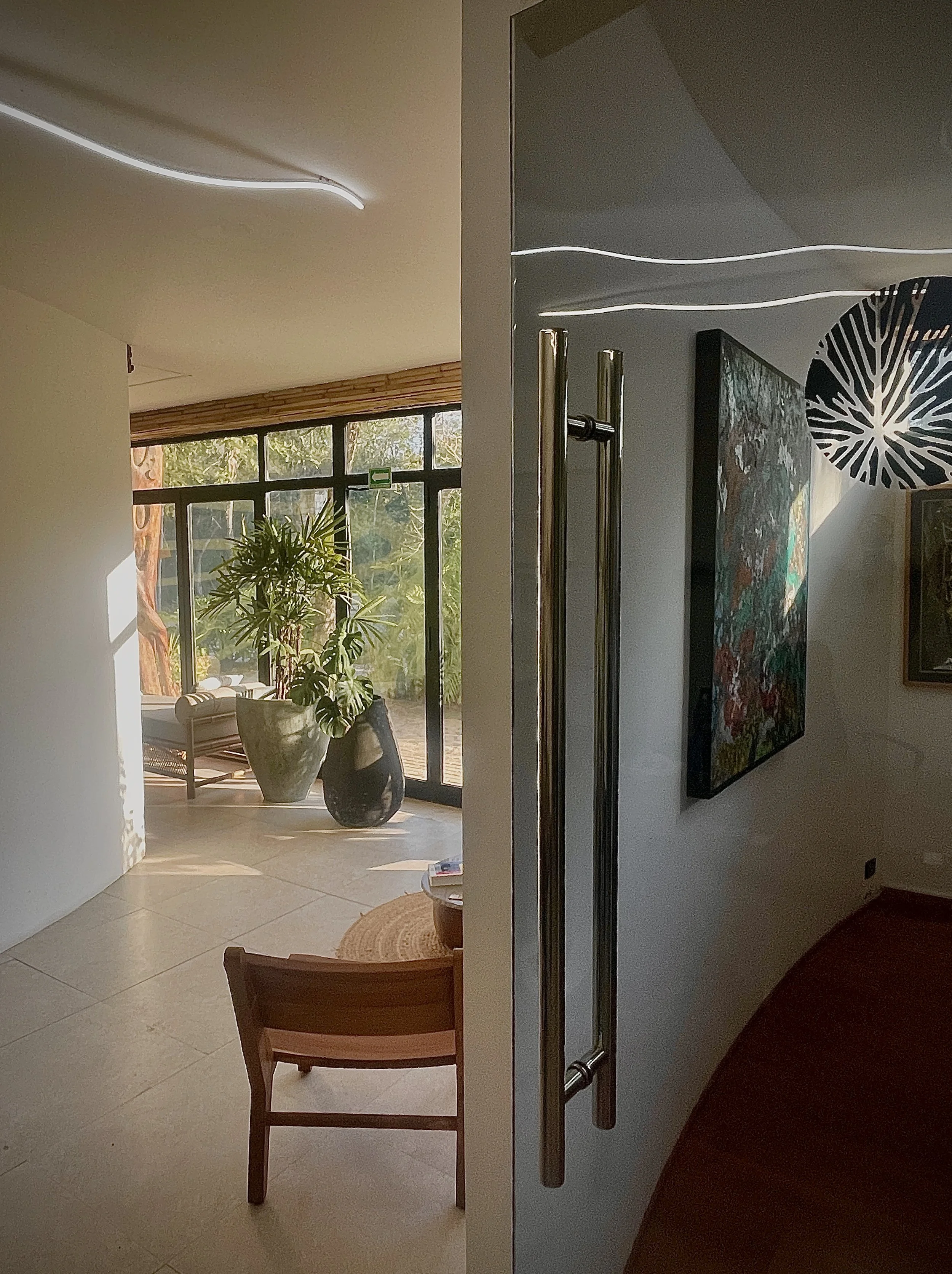 Interior view of a modern space with a door partially open, revealing a sunlit room with large windows, a potted plant, a chair, and artwork on the walls.