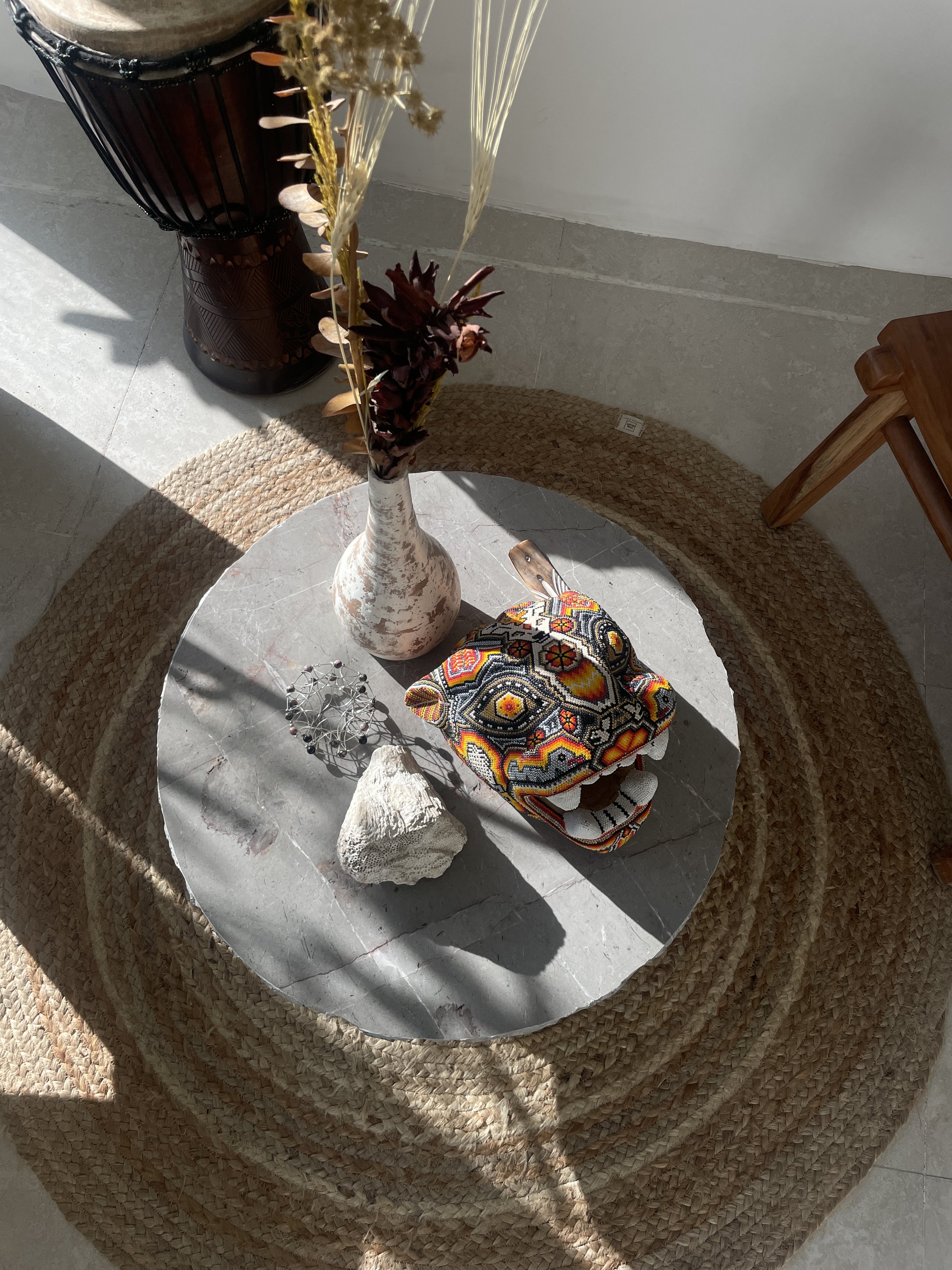 A round tabletop with a gray marble surface, featuring a tall white vase with dried flowers, a carved stone, a colorful embroidered bag with a dinosaur face, and a wire sculpture, all on a woven circular rug. Hypha Inc Marketing Agency 