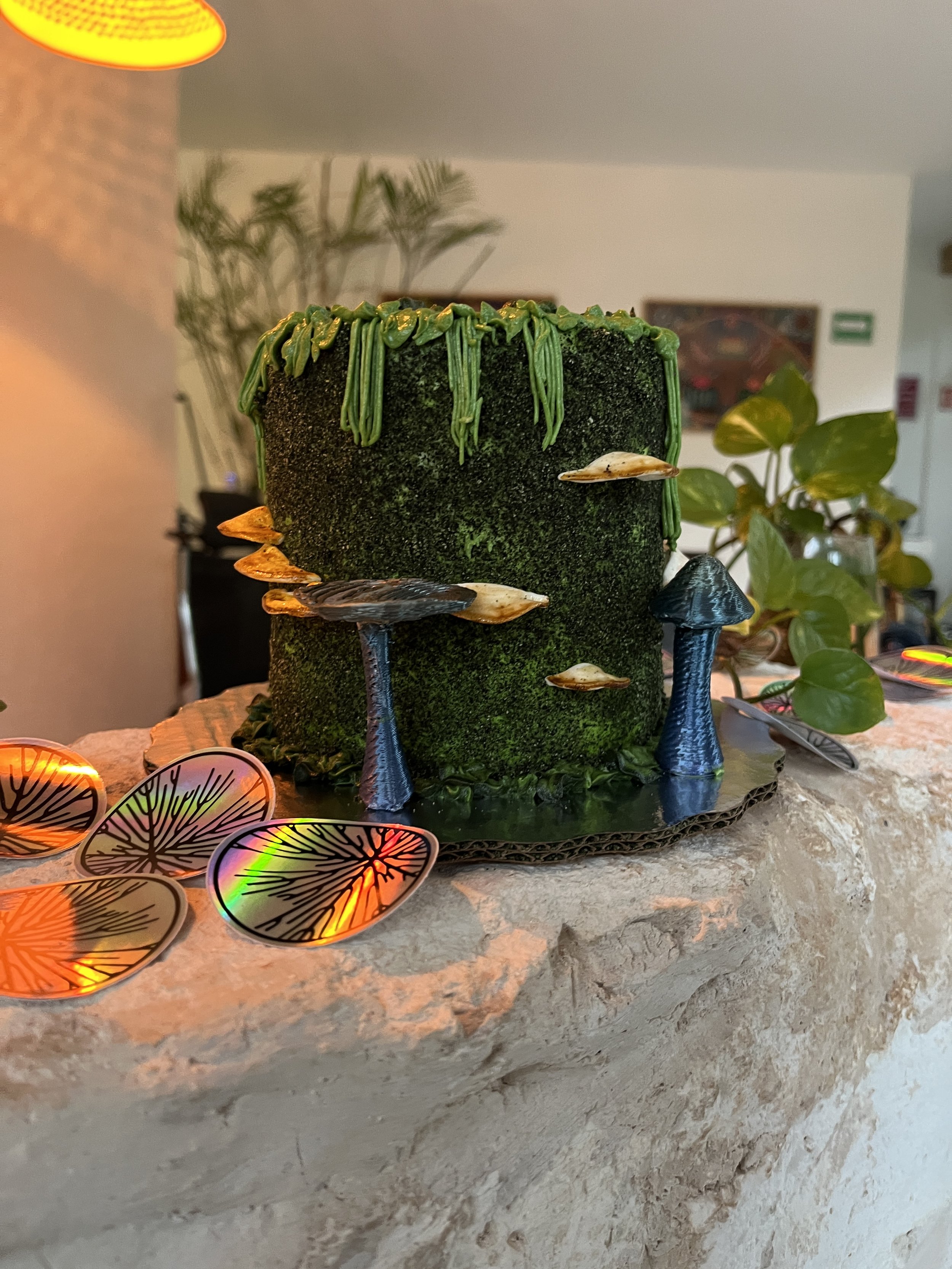 A decorative scene featuring a moss-covered cylindrical object with green dripping details on top, surrounded by small sculpted mushrooms and holographic leaf-shaped confetti on a rock surface, with a blurred background of indoor plants and artwork. 