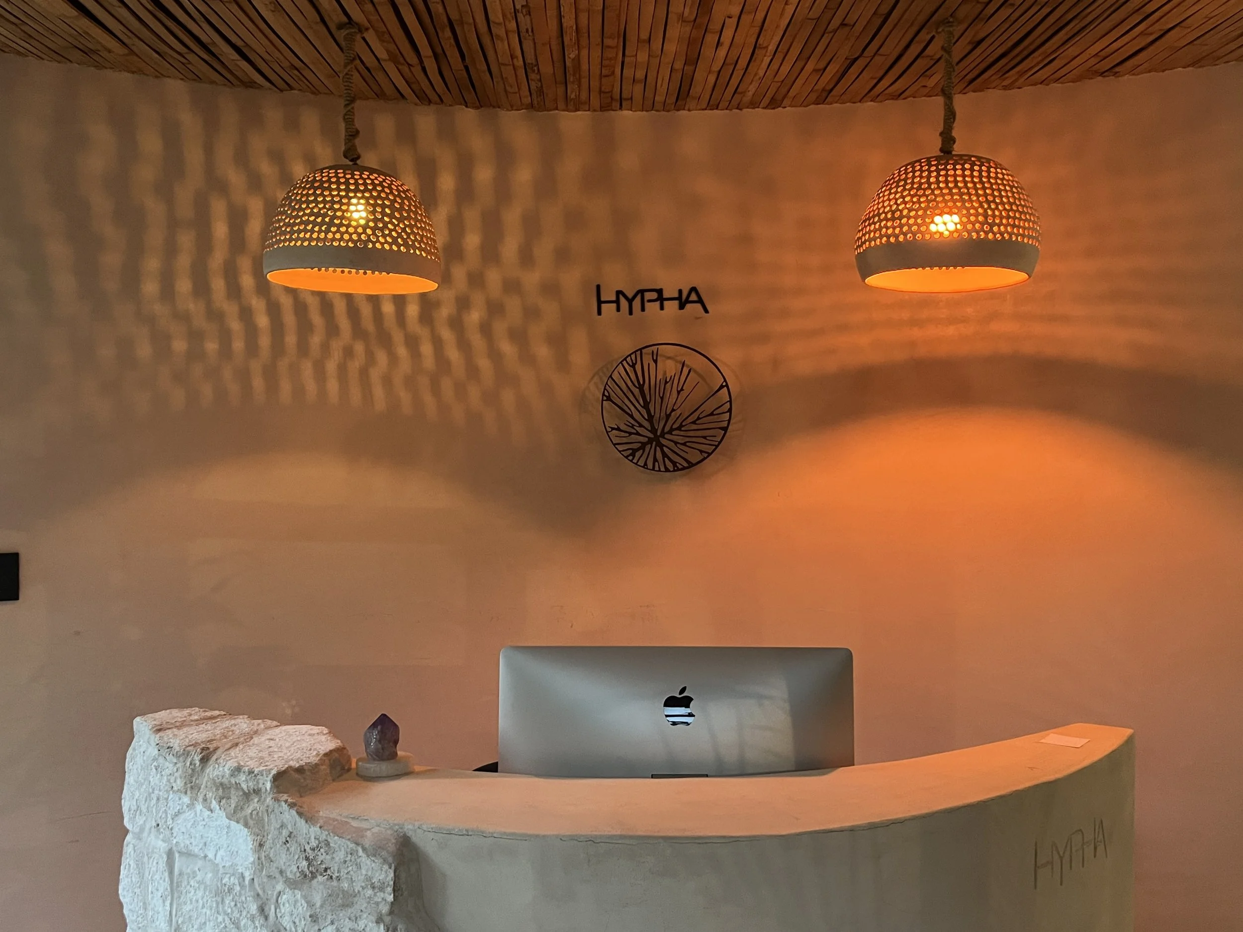 Hypha reception desk with an Apple computer, two woven pendant lights casting warm orange light, a small crystal on stone structure, and branding that reads 'HYPHA' on wall behind the desk with a circle logo and tree design, in a cozy interior space.