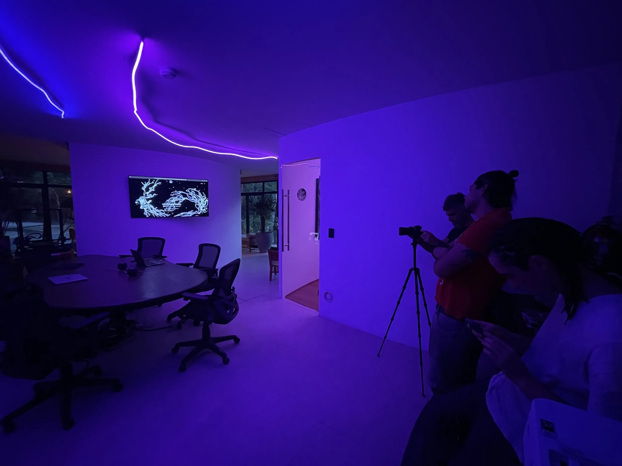 Dimly lit conference room with purple LED lighting, a table with chairs, a wall-mounted monitor displaying digital art, and three people, one with a camera on a tripod, all observing at the side of the room. Hypha Inc Marketing Agency Creative Studio