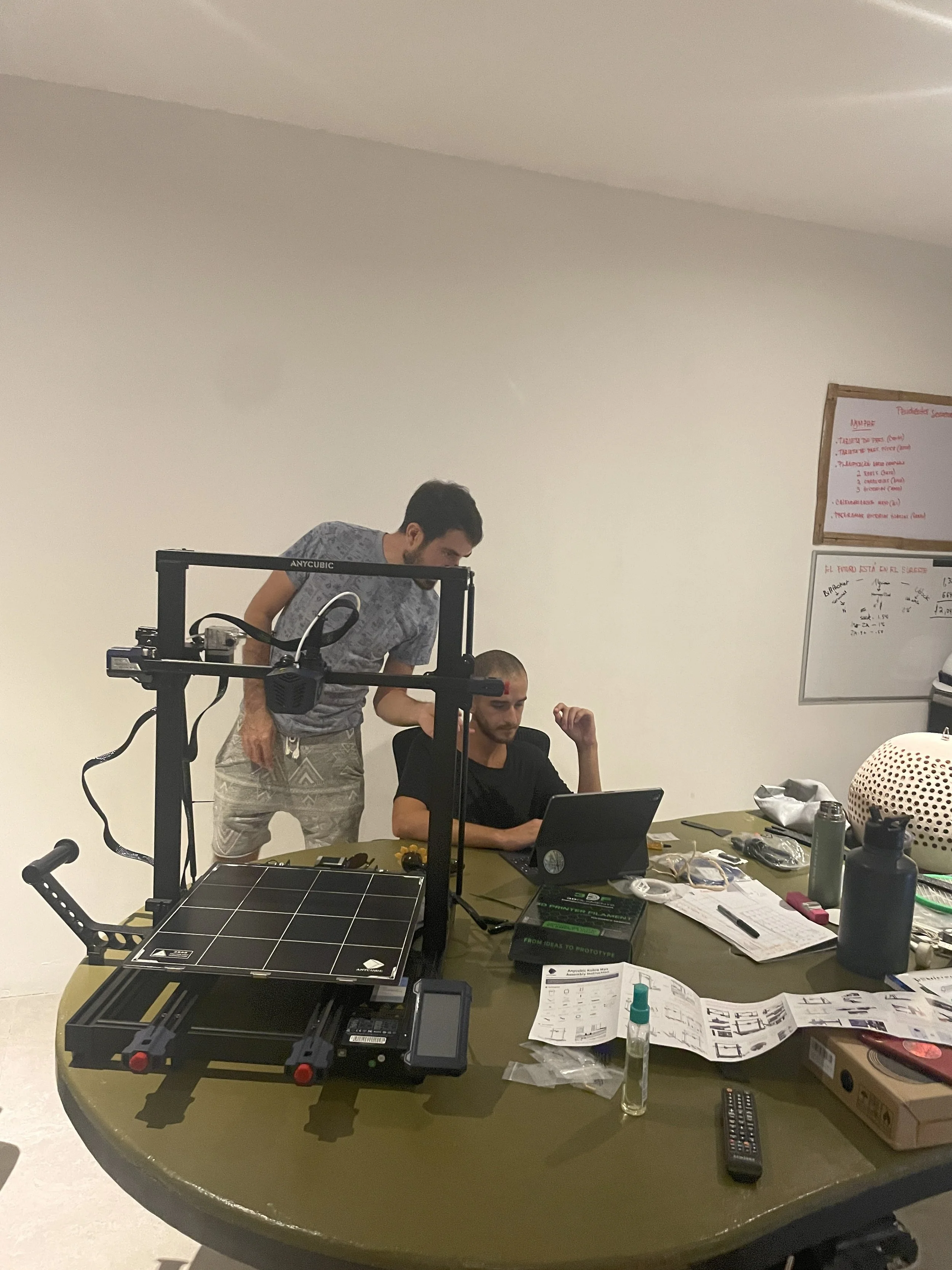 Two men working on a 3D printer at a cluttered table, with a whiteboard in the background. Hypha Inc. Digital Marketing & Advertising Agency, Tulum