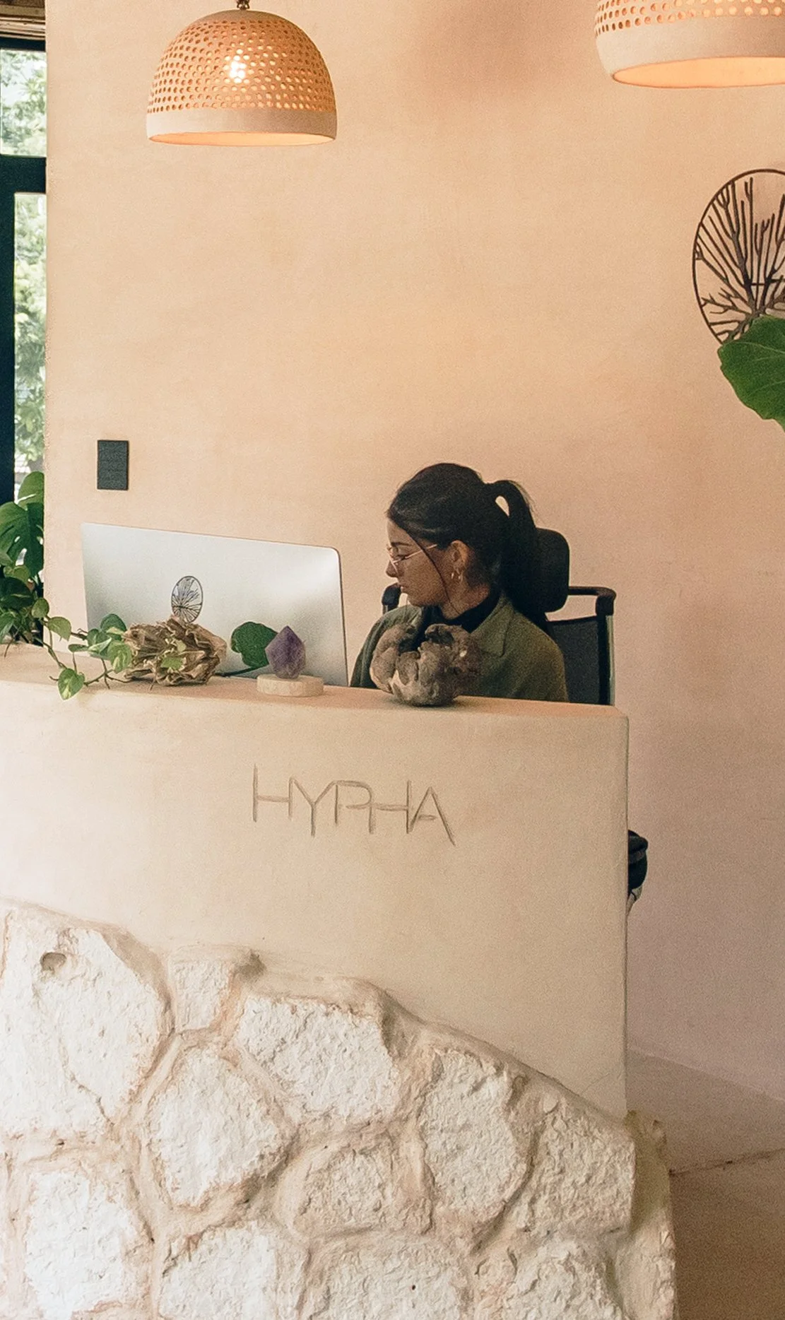 A woman sitting at a reception desk with the word HYPHA engraved on it. She has glasses, dark hair tied back, and is looking at a computer monitor. The reception area has hanging pendant lights, green plants, and decorative stones, with a window show