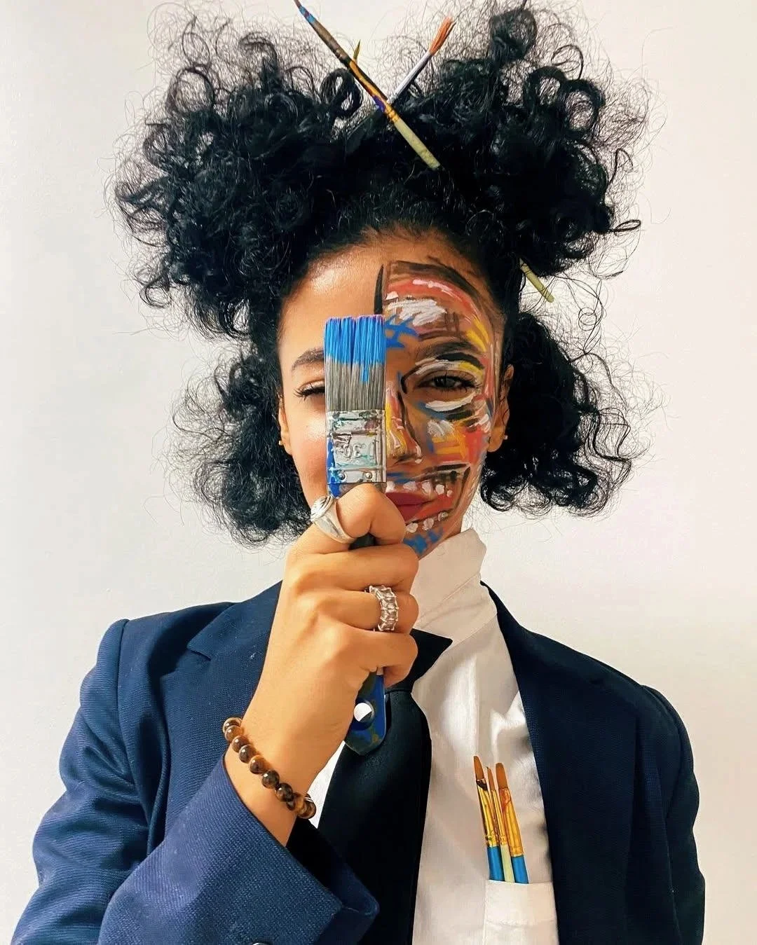 A woman in a suit with a white shirt and black tie holding a paintbrush in front of her face. Her face is painted with abstract colorful designs, and her hair is styled in curly updo with brushes stuck in it.