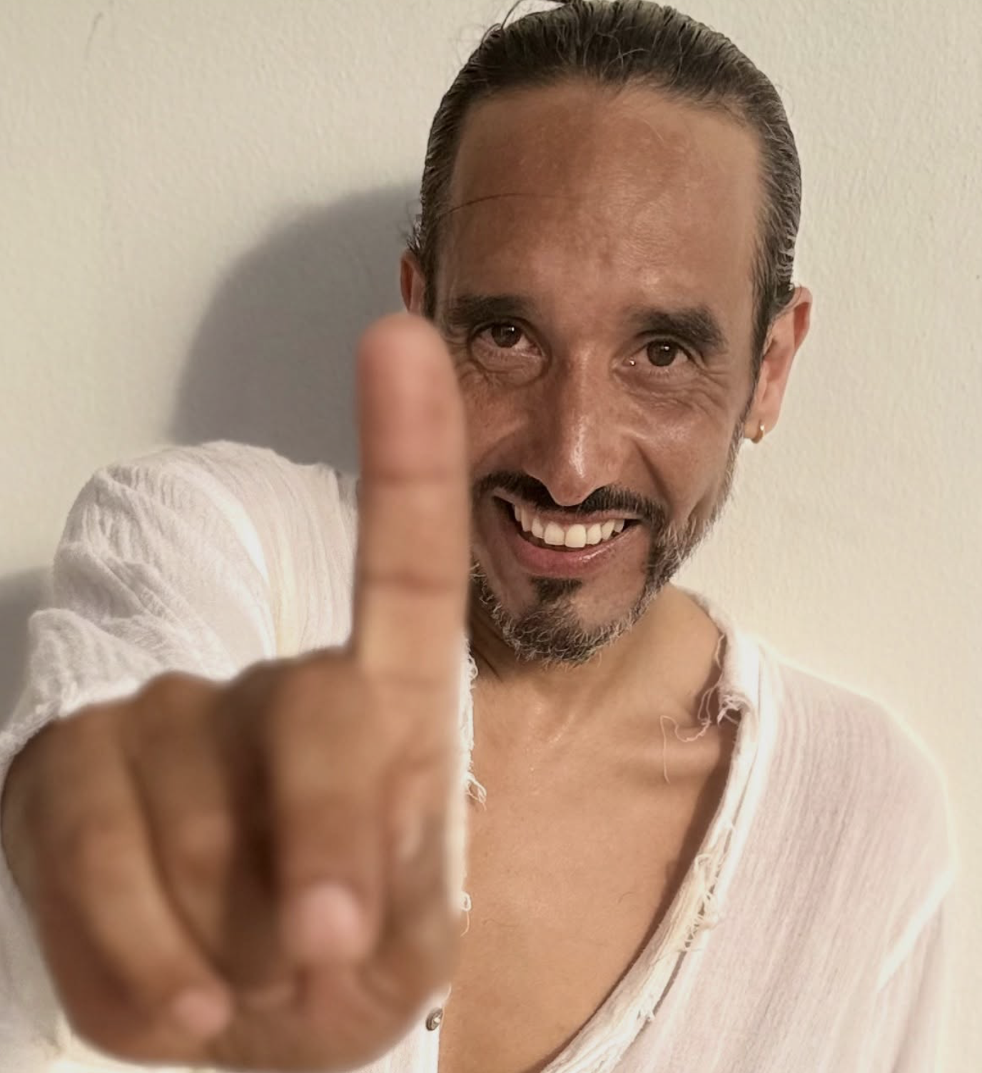 Jose Fernando Sanchez, smiling with dark hair and a beard, wearing a wet white shirt, extends his middle finger towards the camera, standing against a plain wall.