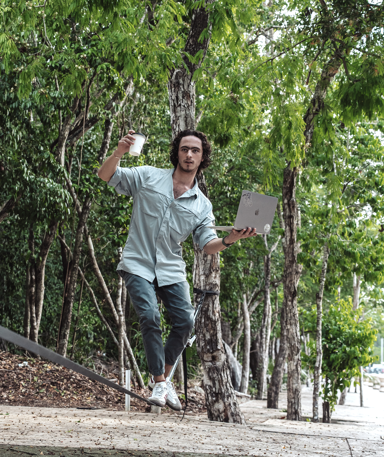 A young man with curly hair balancing on a slackline in a wooded park, holding a coffee cup in one hand and a laptop in the other. Pato Herrera Hypha Inc.
