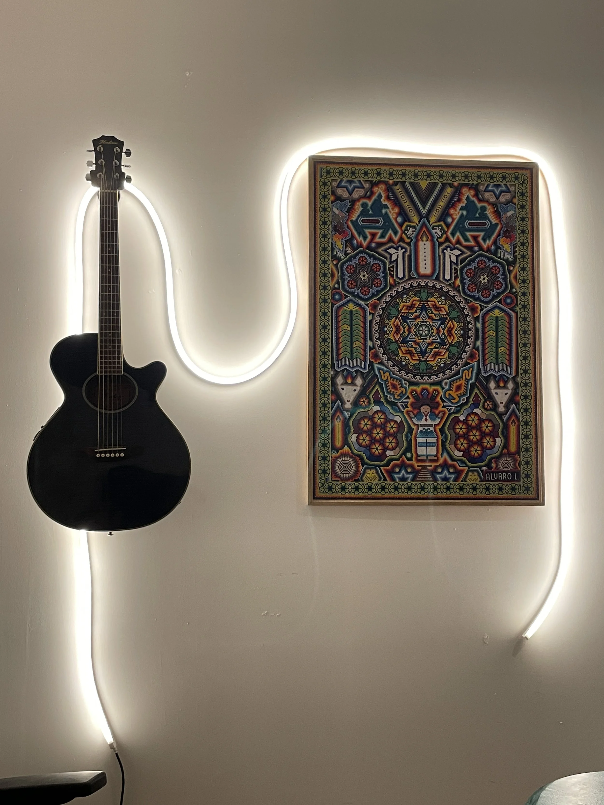 Decorative wall art with neon light accents, featuring a black acoustic guitar mounted on the wall and a colorful, intricate Huichol artwork surrounded by neon lights. Hypha Inc Marketing Agency and Creative Studio Lab, Tulum.