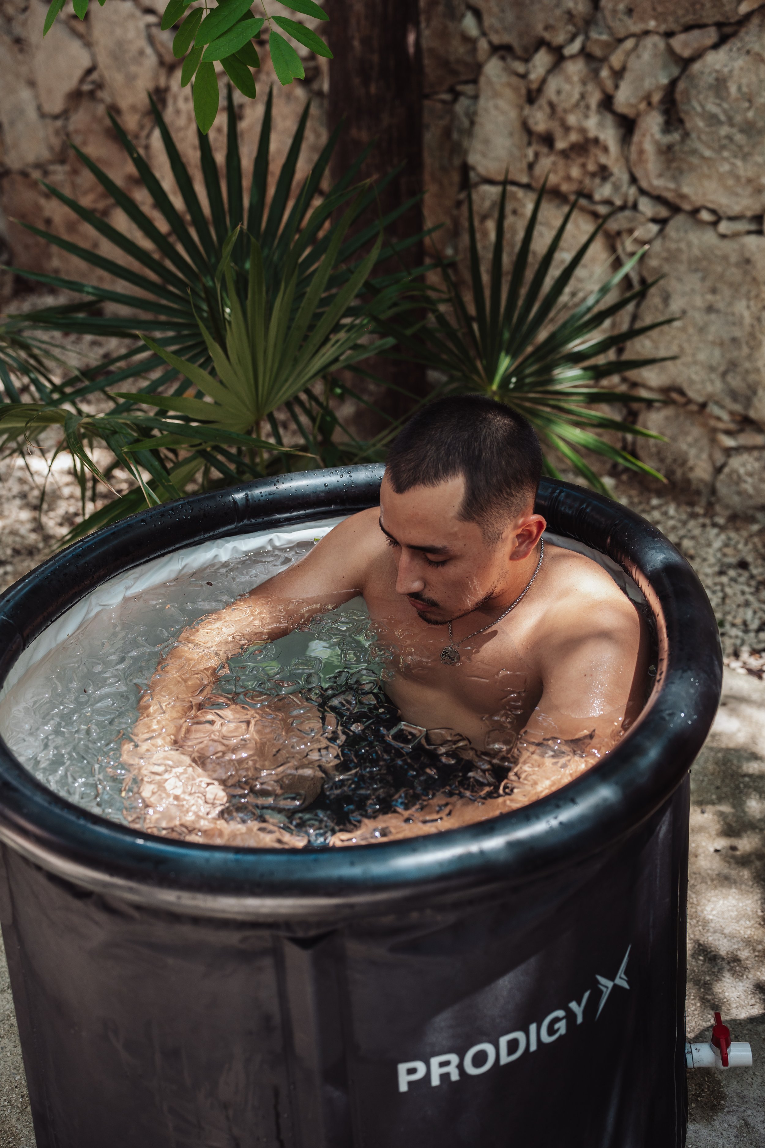 A shirtless man sitting in a black Prodigy brand hot tub outdoors among green plants and rocks.