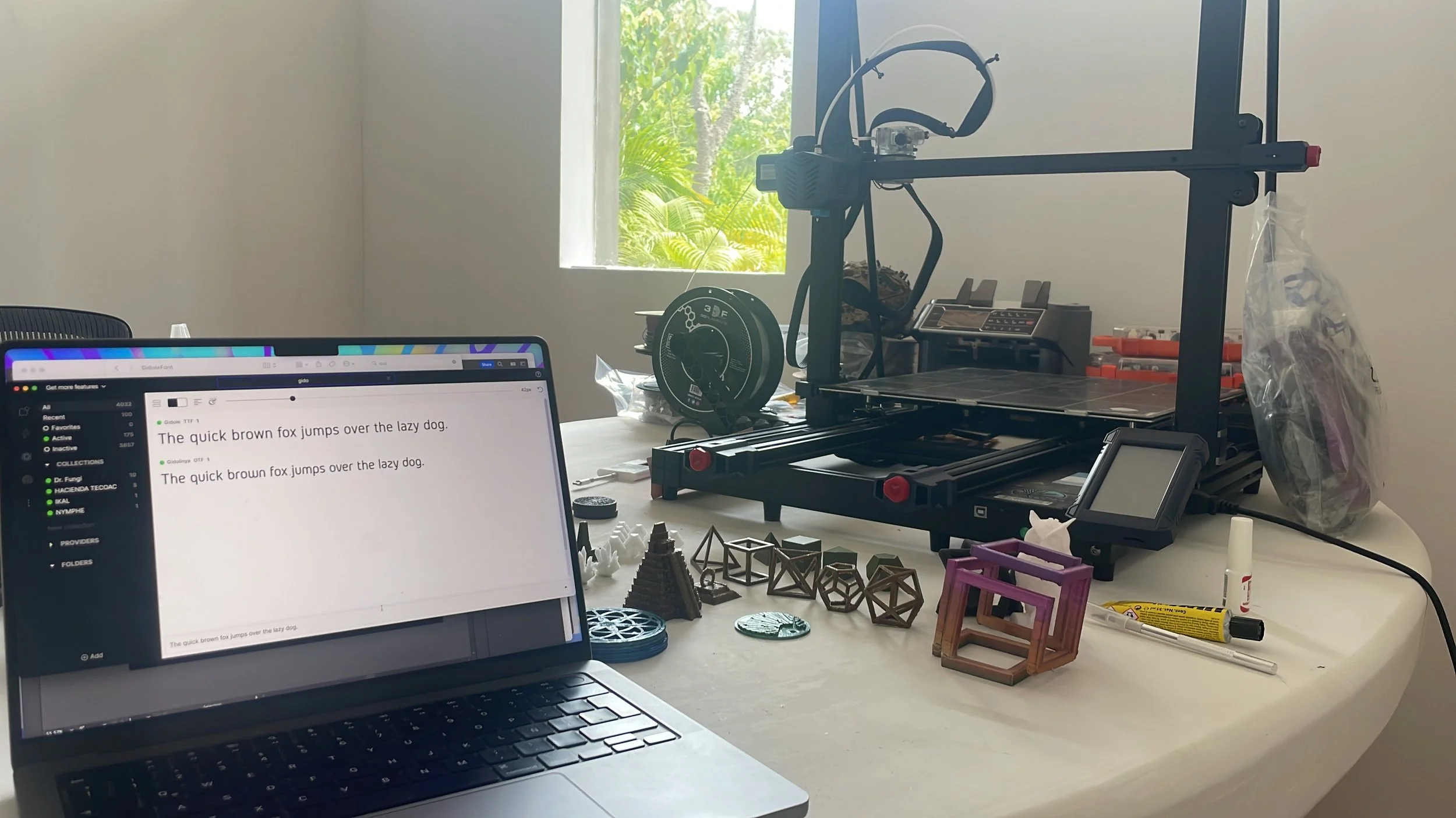A room with a desk holding a laptop, 3D printer, various 3D printed objects, tools, and supplies, with a window showing green trees outside. Hypha Inc Marketing Agency and Creative Studio Lab, Tulum.