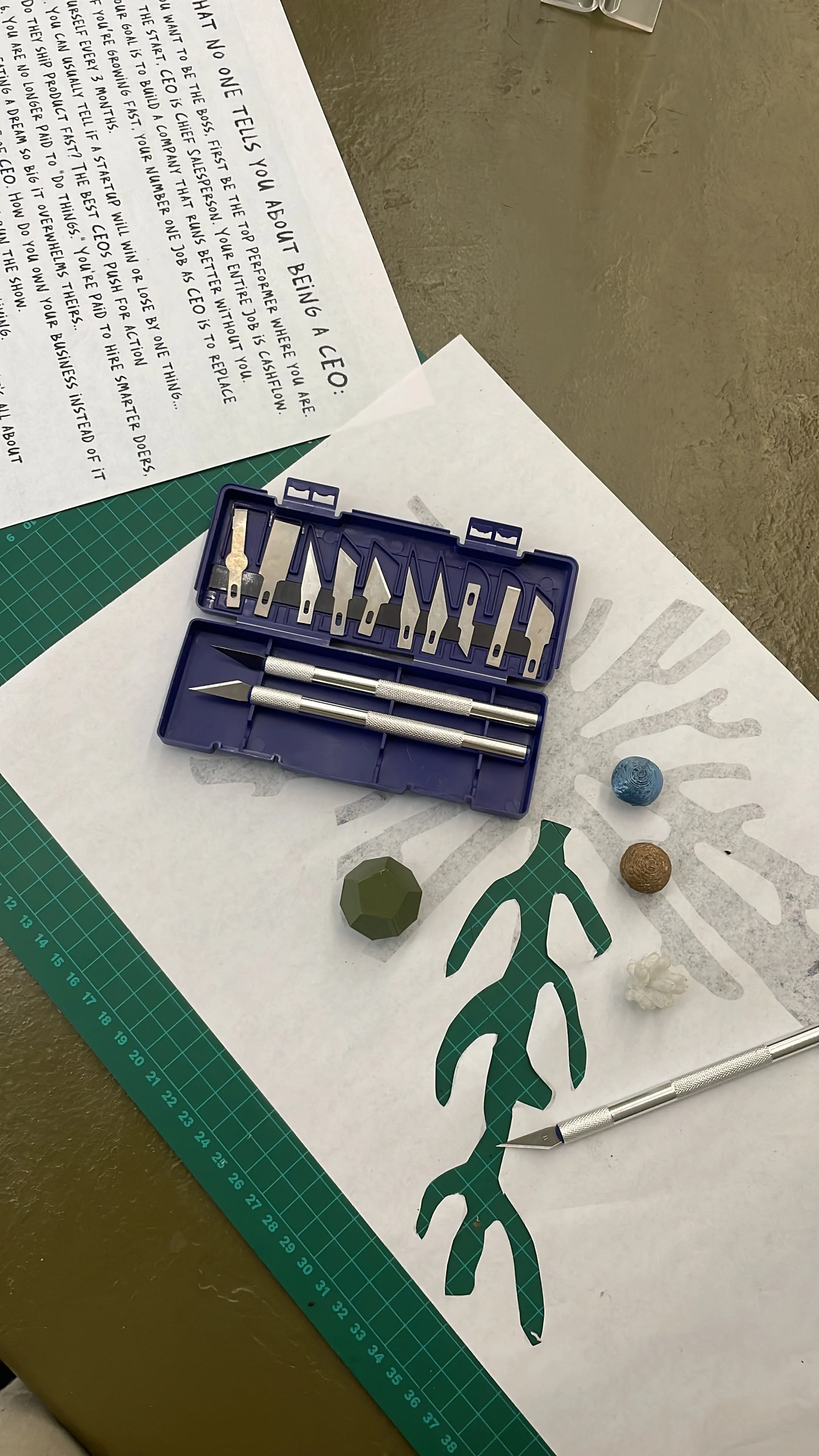 A craft workspace featuring a cutting mat, a paper cutout of a leafy branch, a set of precision craft knives, four small decorative stones in blue, green, brown, and white, and a printed sheet of text. Hypha Inc Marketing Agency and Creative Studio