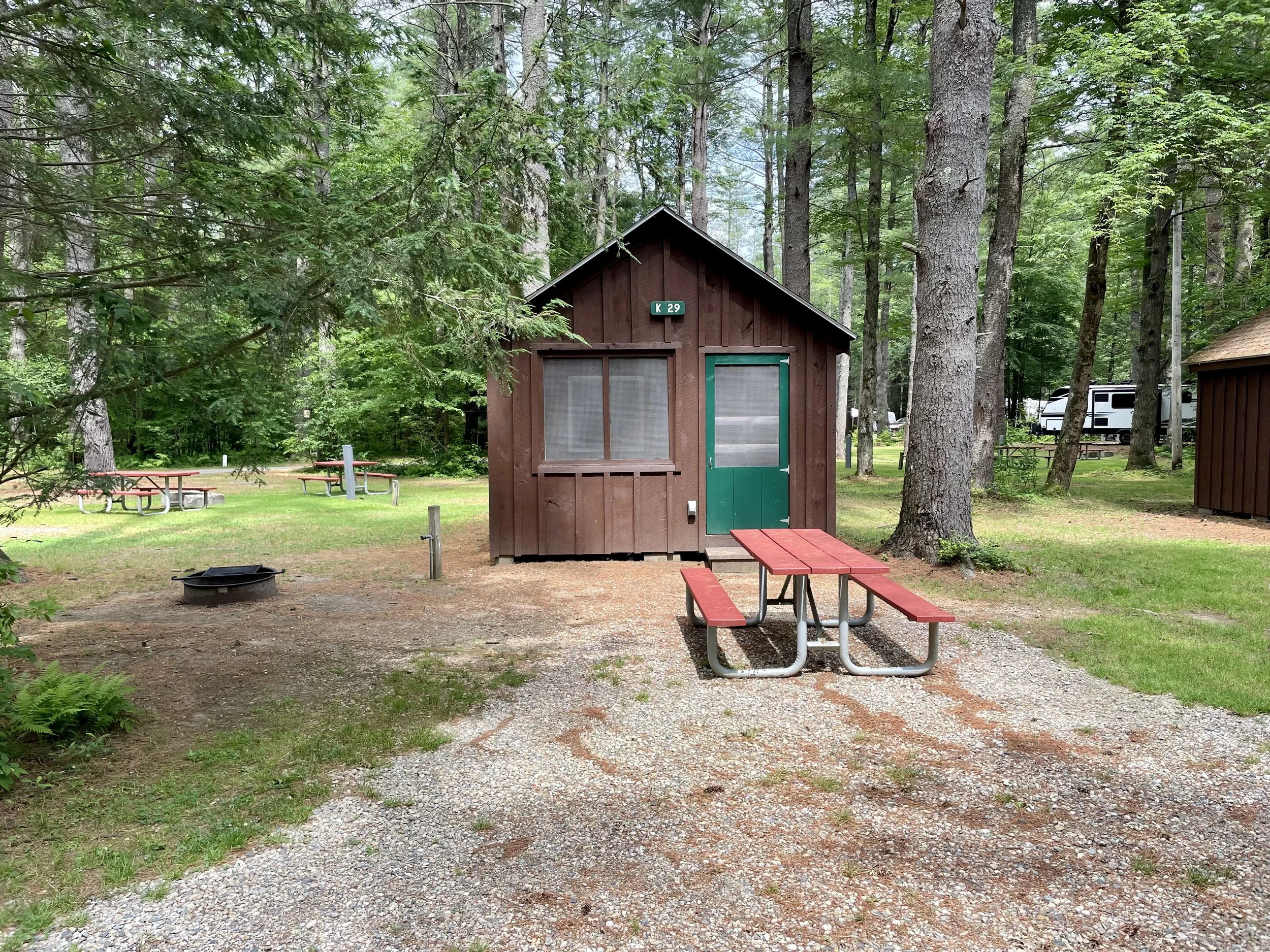 A small wooden cabin in a wooded campground with a green door and a window, surrounded by trees and grass, with a red picnic table in front and additional picnic tables in the background.