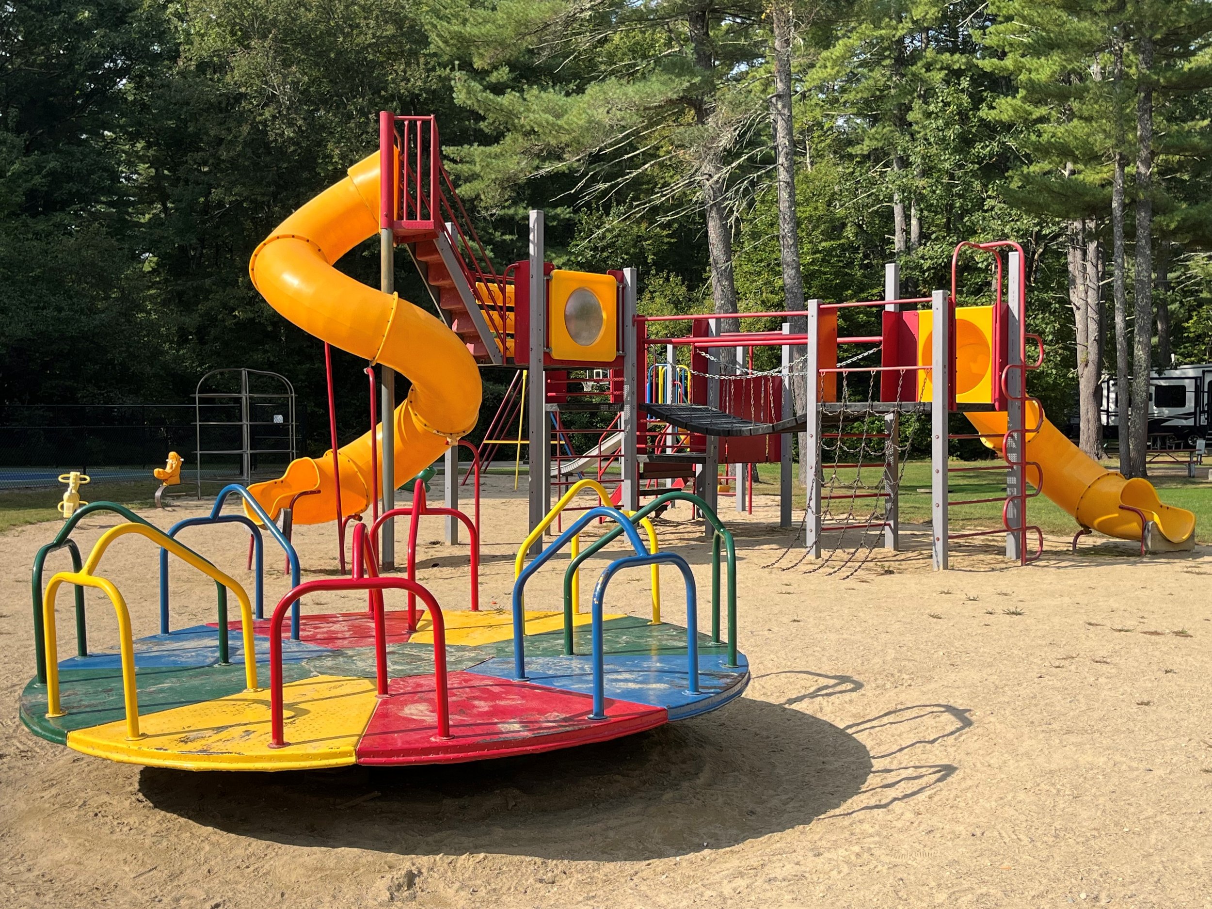 A playground with a red-roofed pavilion, a multicolored climbing structure with slides, and swings, surrounded by trees and a clear blue sky.